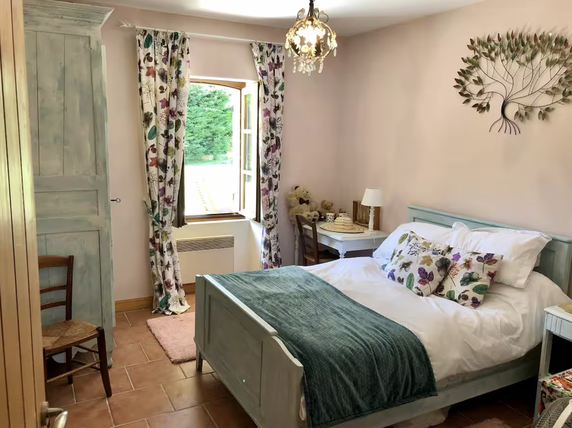 Cozy bedroom with pastel-colored furniture, floral curtains, a bed with white sheets and floral pillows, a window with open curtains showing greenery outside, a chandelier hanging from the ceiling, and decorative wall art.