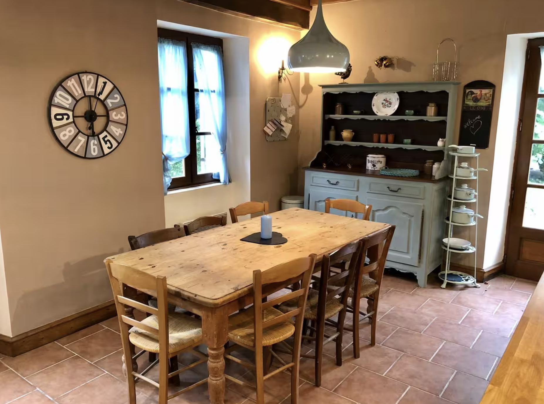 A rustic dining room with a wooden table, six chairs, a large wall clock, a window with blue curtains, a vintage hutch with dishes and decor, and a small shelf with plates. There is a candle on the table and soft lighting.