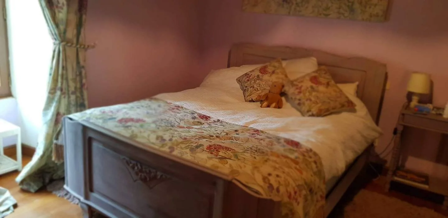 A cozy bedroom with a wooden bed featuring floral bedding and pillows, a nightstand with a lamp, and light-colored curtains next to a window.
