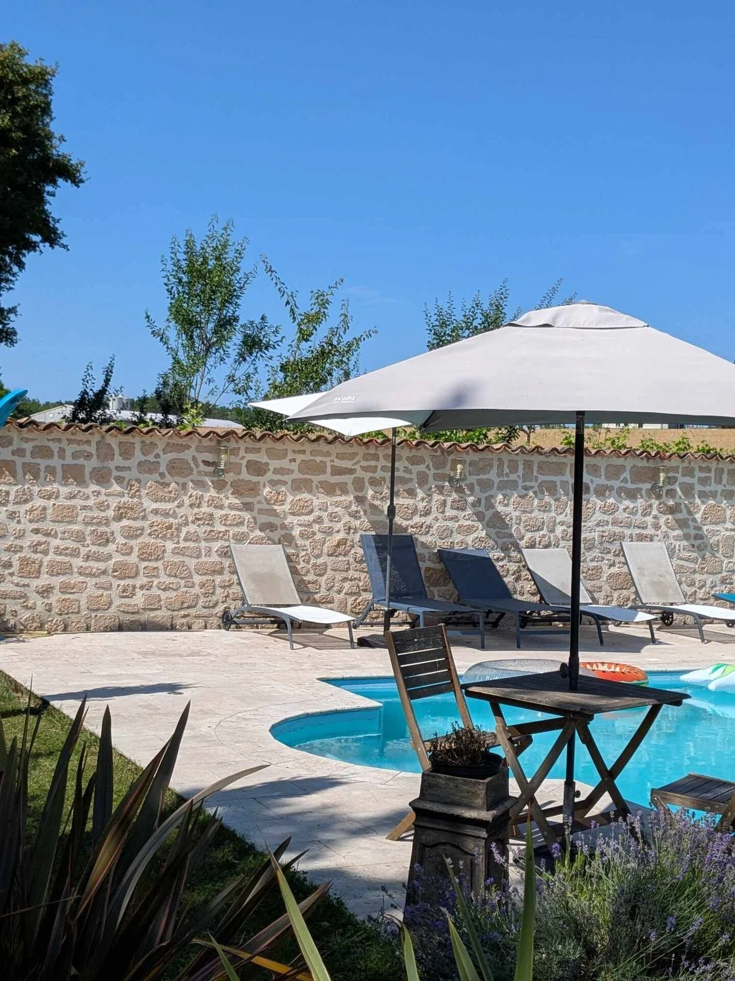 Outdoor swimming pool with lounge chairs, umbrellas, and a stone wall in the background under a clear blue sky.