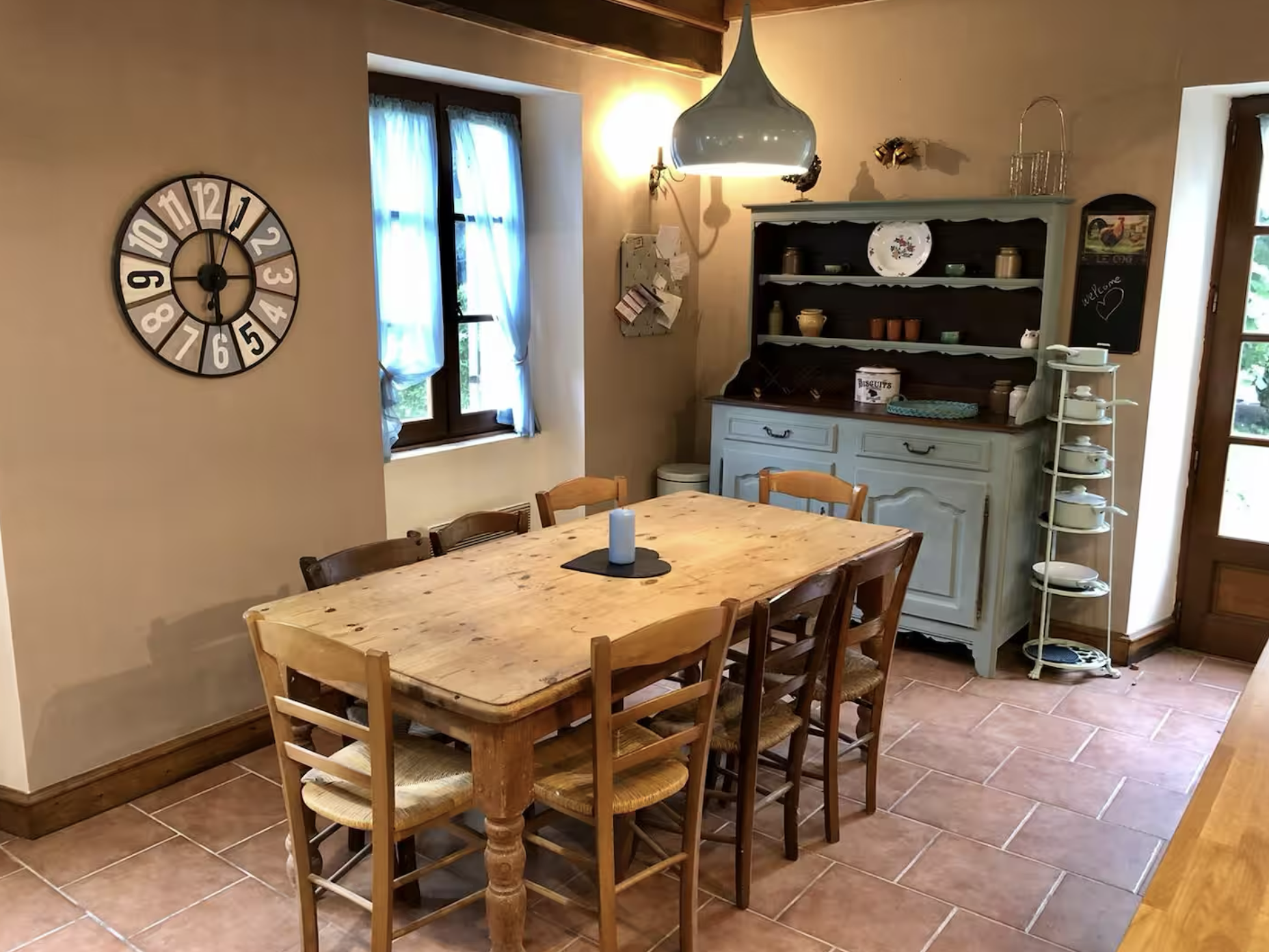 A cozy dining room with a wooden table and six chairs, a vintage-style hutch with dishes, a wall clock, two windows with blue curtains, a white towel rack with plates, and various wall decorations in a warm, rustic style.