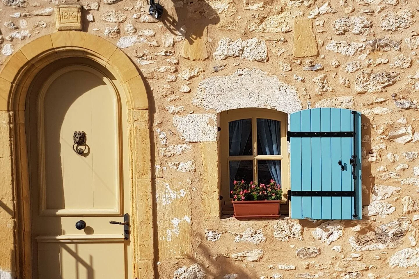 A beige door with a lion-shaped door knocker and a black doorknob, next to a window with an open blue shutter, a flower box with pink flowers, and a stone wall.