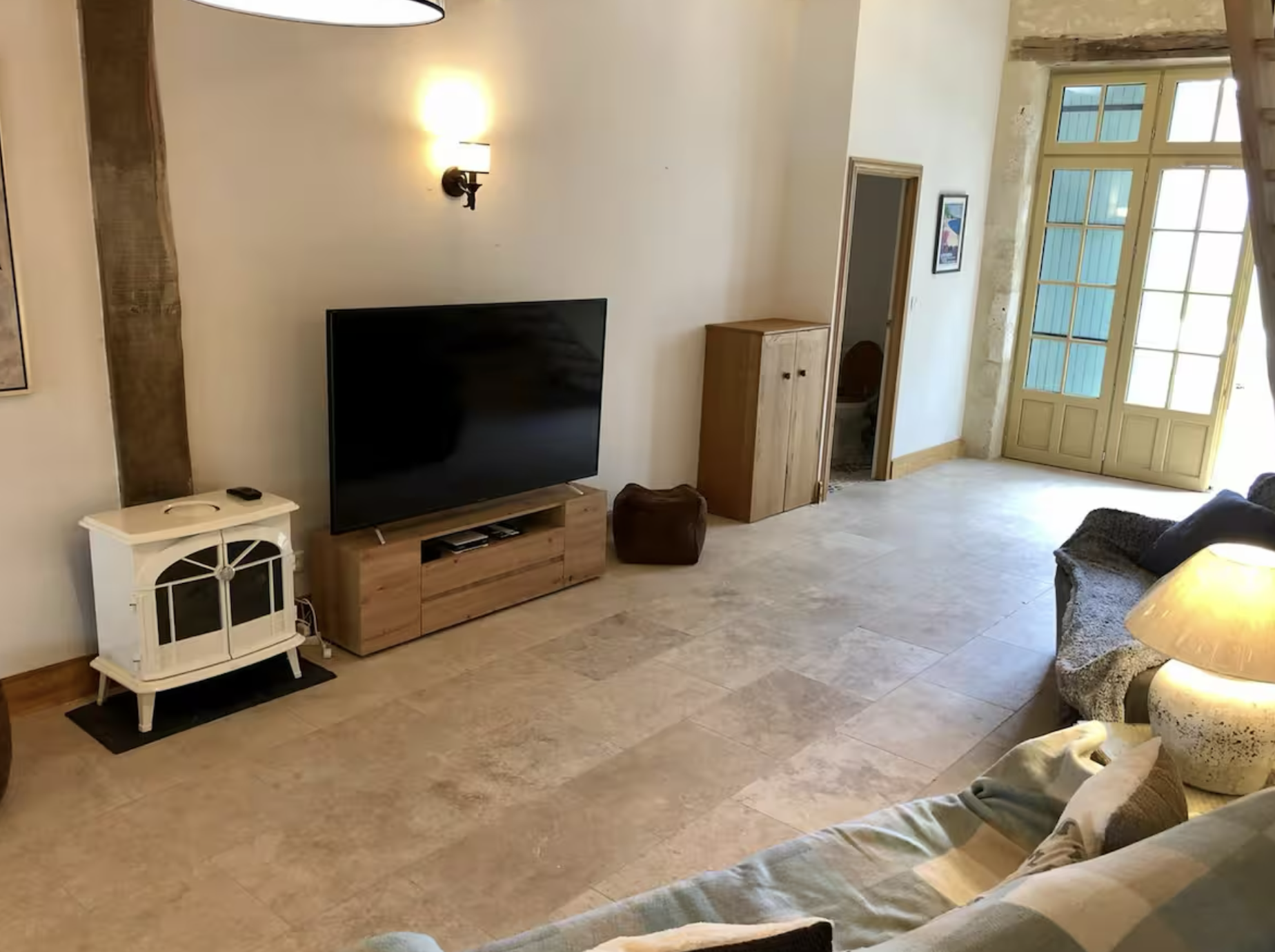 Living room with a flat-screen TV on a wooden stand, a small white cabinet, a brown pouf, a wooden cabinet, a sofa with a pillow, a lamp, and a large glass door leading outside.