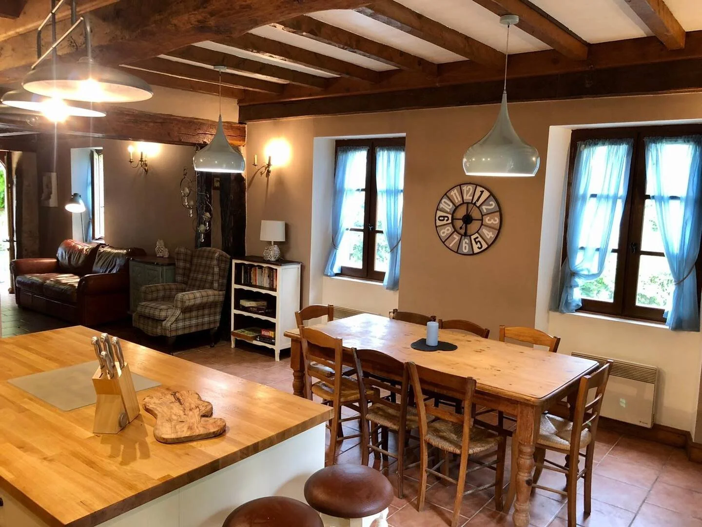 Interior of a rustic home with a wooden dining table and chairs, a sitting area with a leather sofa and plaid armchair, windows with blue curtains, and warm lighting.