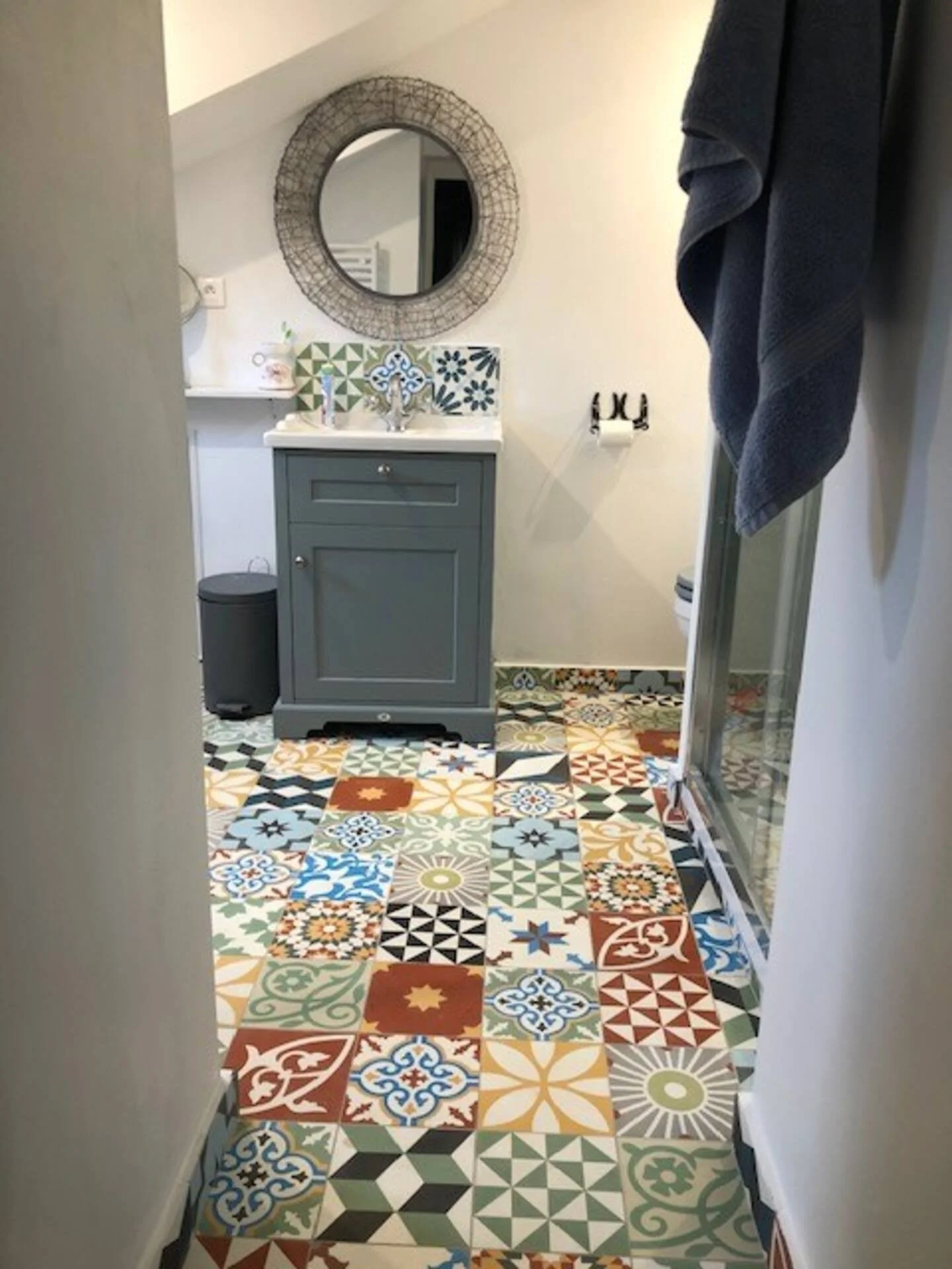 A small bathroom with colorful patchwork tile flooring, a blue vanity with a white countertop and blue-patterned backsplash tiles, a round mirror with a wire frame, a towel hanging on the right, and a Trash can on the left.
