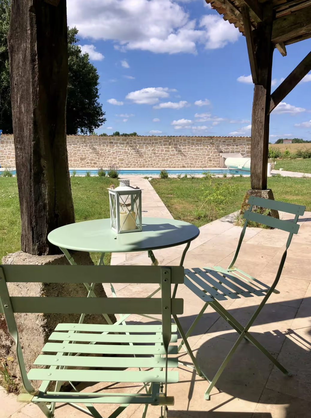A peaceful backyard patio with a small round table and two matching chairs under a wooden pergola. A lantern with a candle sits on the table. In the background, there's a swimming pool with a stone pathway leading to it, a stone wall, and a bright bl
