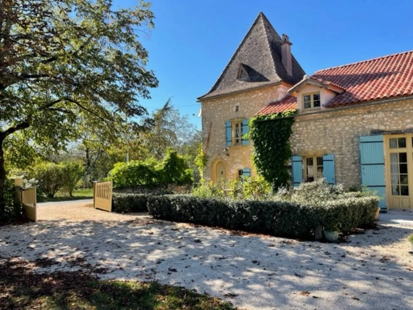 Stone house with red tiled roof, blue shutters, and ivy growing on the wall, surrounded by trees and bushes under a clear blue sky.