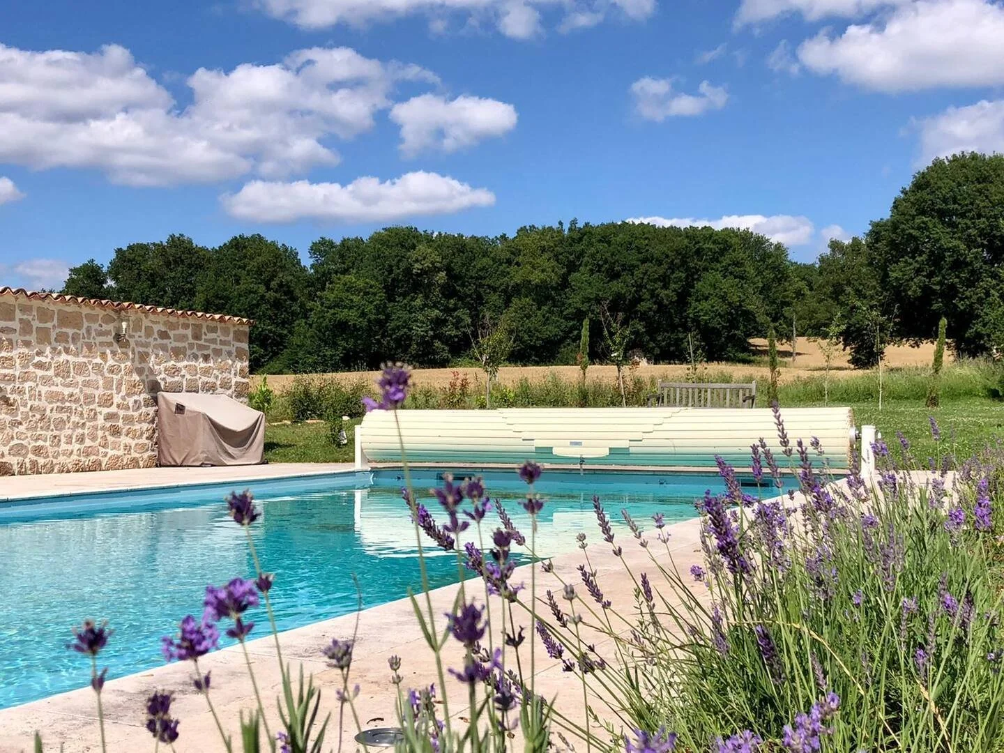 Outdoor swimming pool with purple flowers in the foreground, stone building to the left, and a rolled-up pool cover in the background, surrounded by green trees and a blue sky with clouds.