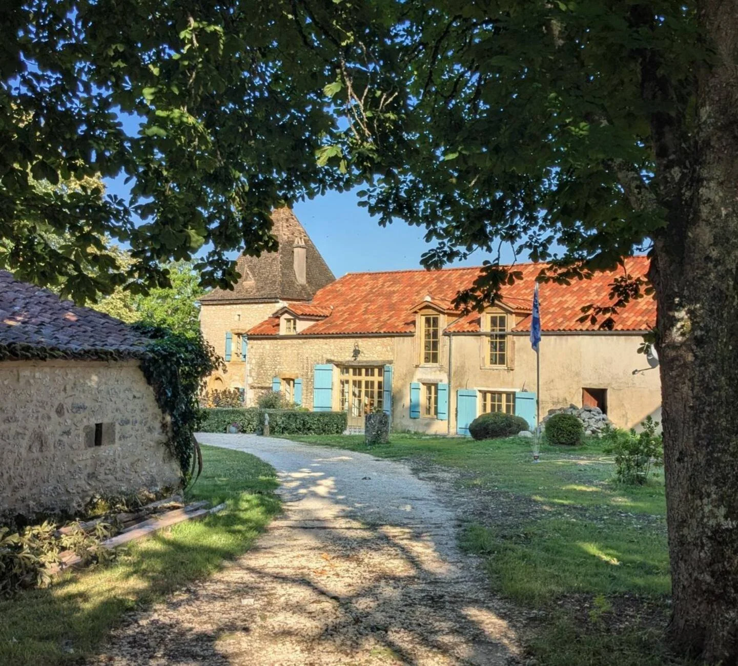 A rustic stone house with blue window shutters and a red tile roof, surrounded by a gravel pathway, trees, and a flagpole in a garden setting.