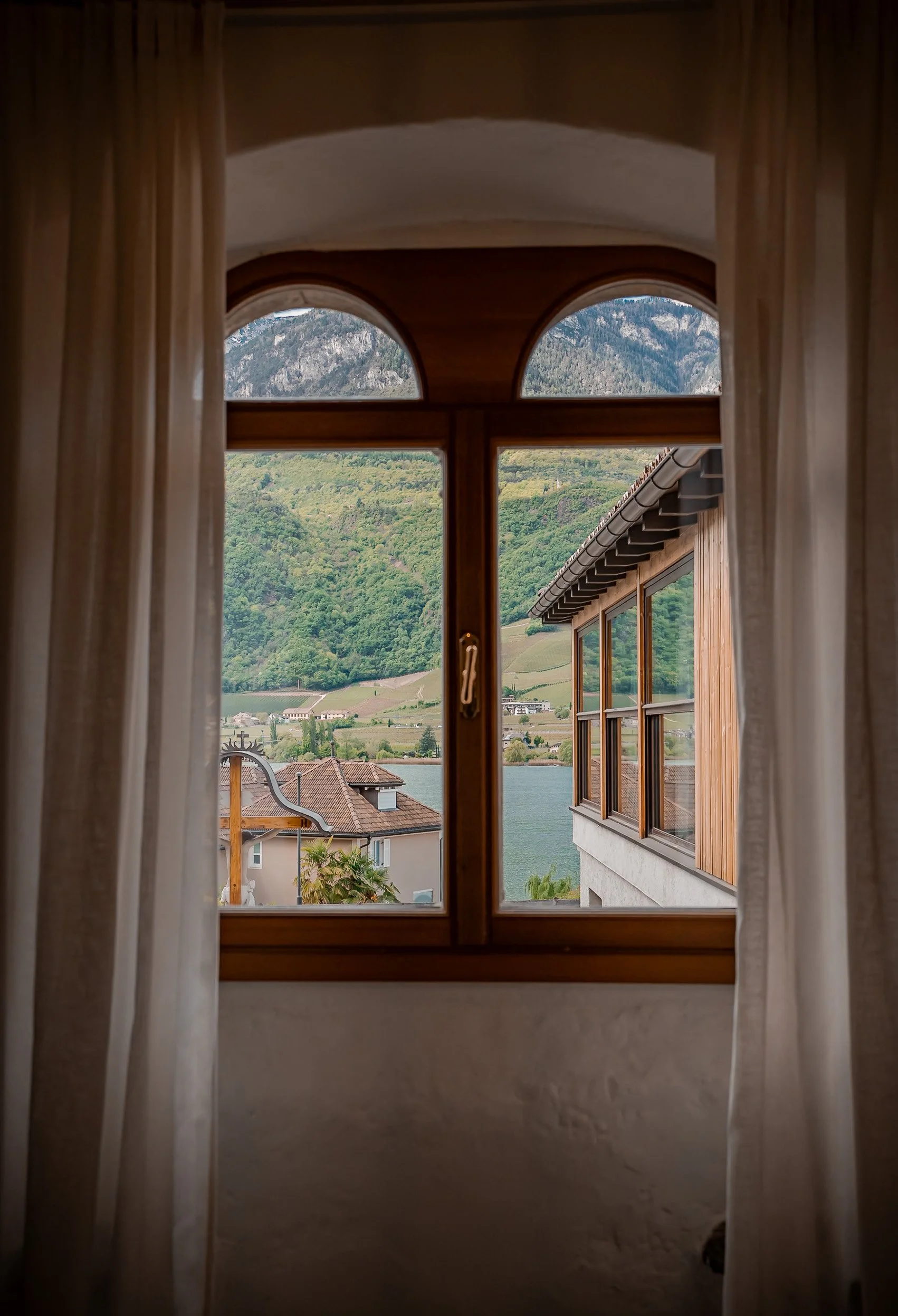 Vista dal finestrino di una casa, con montagne, un lago, edifici e vegetazione circostante.
Blick aus dem Fenster eines Hauses mit Bergen, einem See, Gebäuden und umliegender Vegetation – mit Sicht auf den Kalterer See.