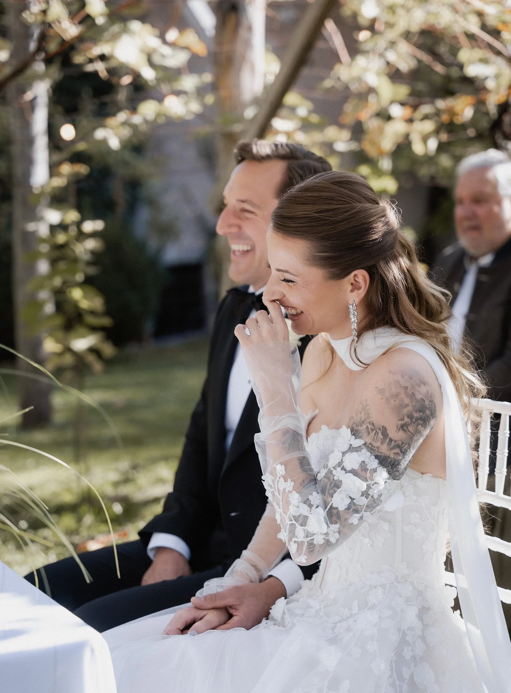 Ein glückliches Brautpaar bei einer Hochzeit im Freien, sitzend Hand in Hand, lachend und in einem festlichen Kleid und einem Anzug, umgeben von Natur und Dekorationen.