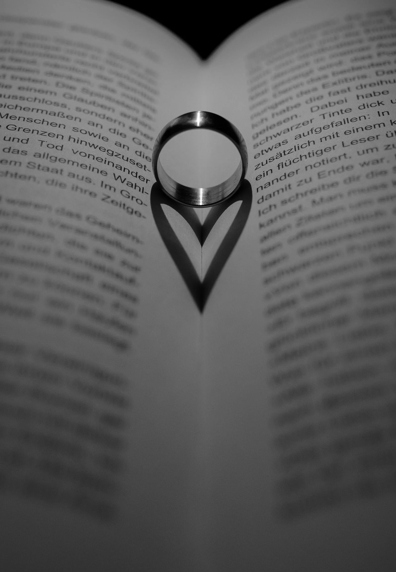 Un anello posto tra le pagine di un libro, creando l'ombra di un cuore sul testo.
Ein Ring liegt zwischen den Seiten eines Buches und wirft einen herzförmigen Schatten auf den Text.