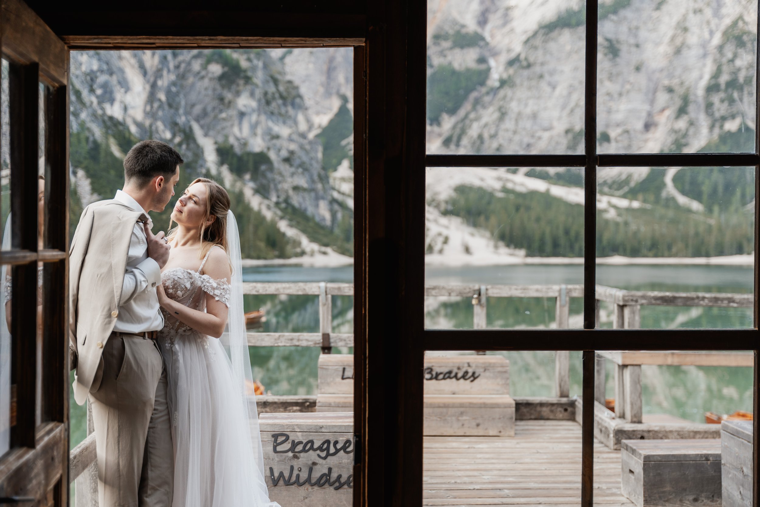 Verliebtes Hochzeitspaar schaut sich an, hinter einer Fensteröffnung mit dunklem Holzrahmen, mit Berglandschaft und Wasser im Hintergrund.