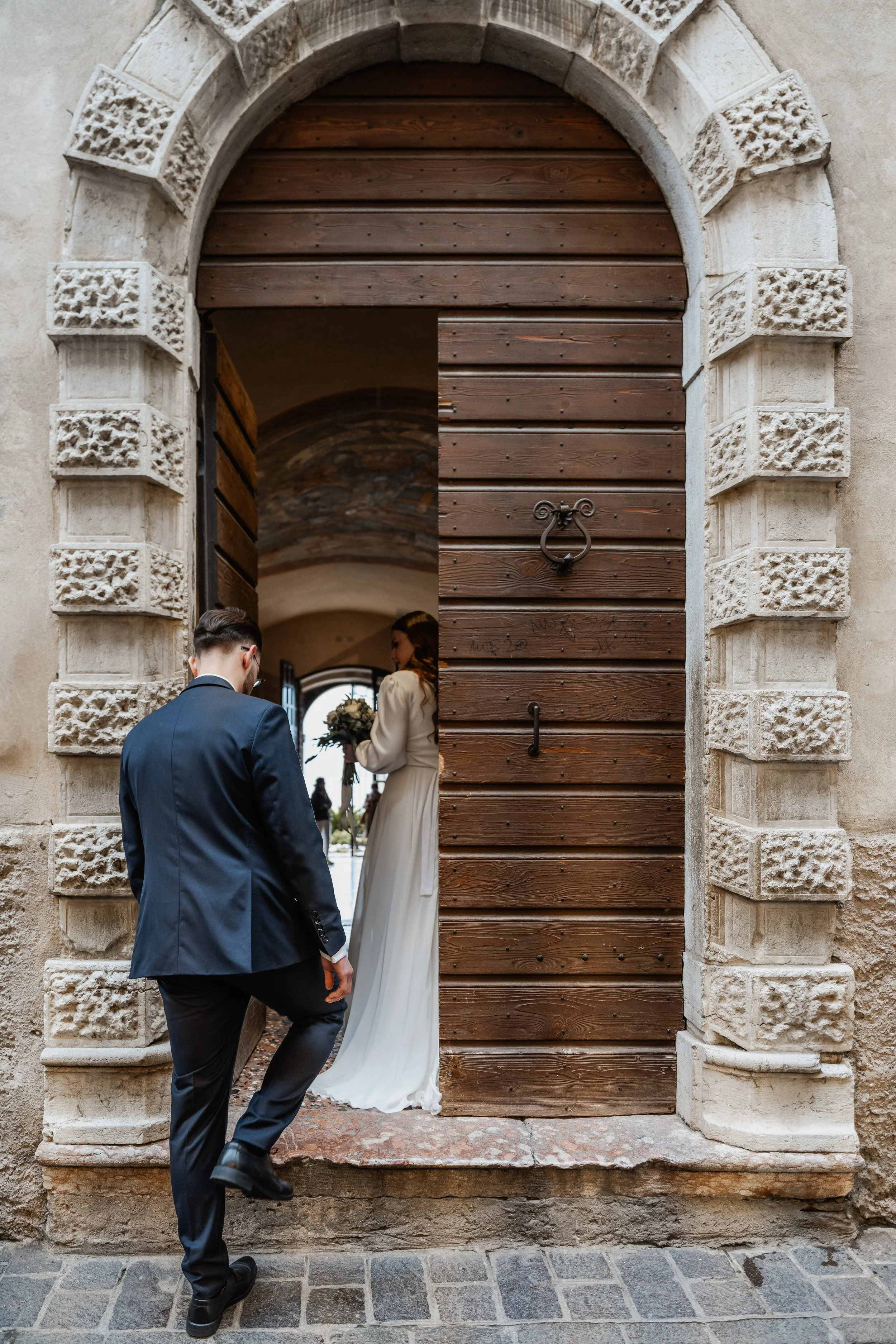 Ein Mann in einem dunklen Anzug mit Brille steht vor einem Holztor, durch das eine Frau in einem weißen Brautkleid mit Blumenstrauß sichtbar ist, während sie in eine Kirche eintreten.