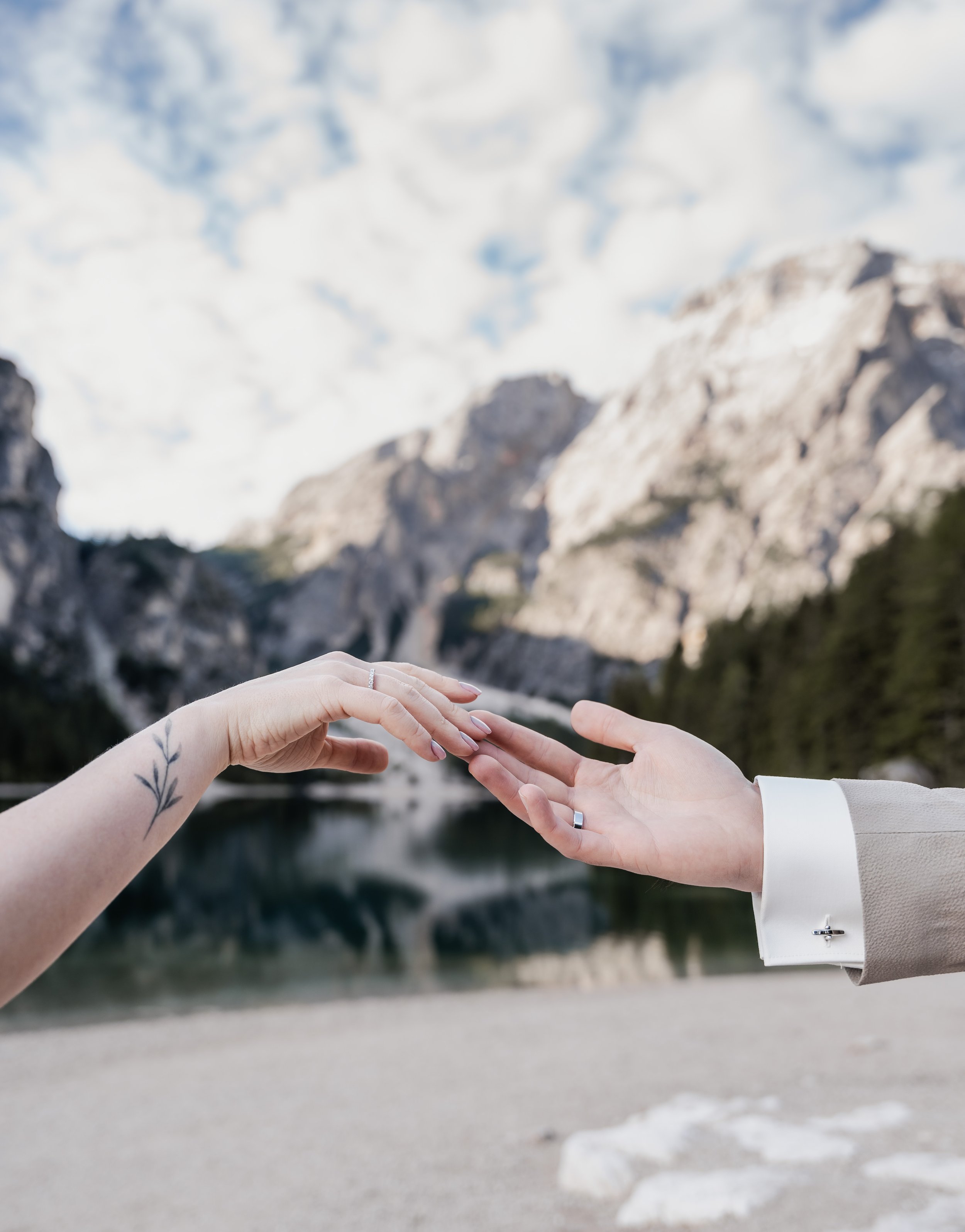 Zwei Hände, eine mit einem Ehering, berühren sich vor einem Bergsee mit Bergen im Hintergrund.