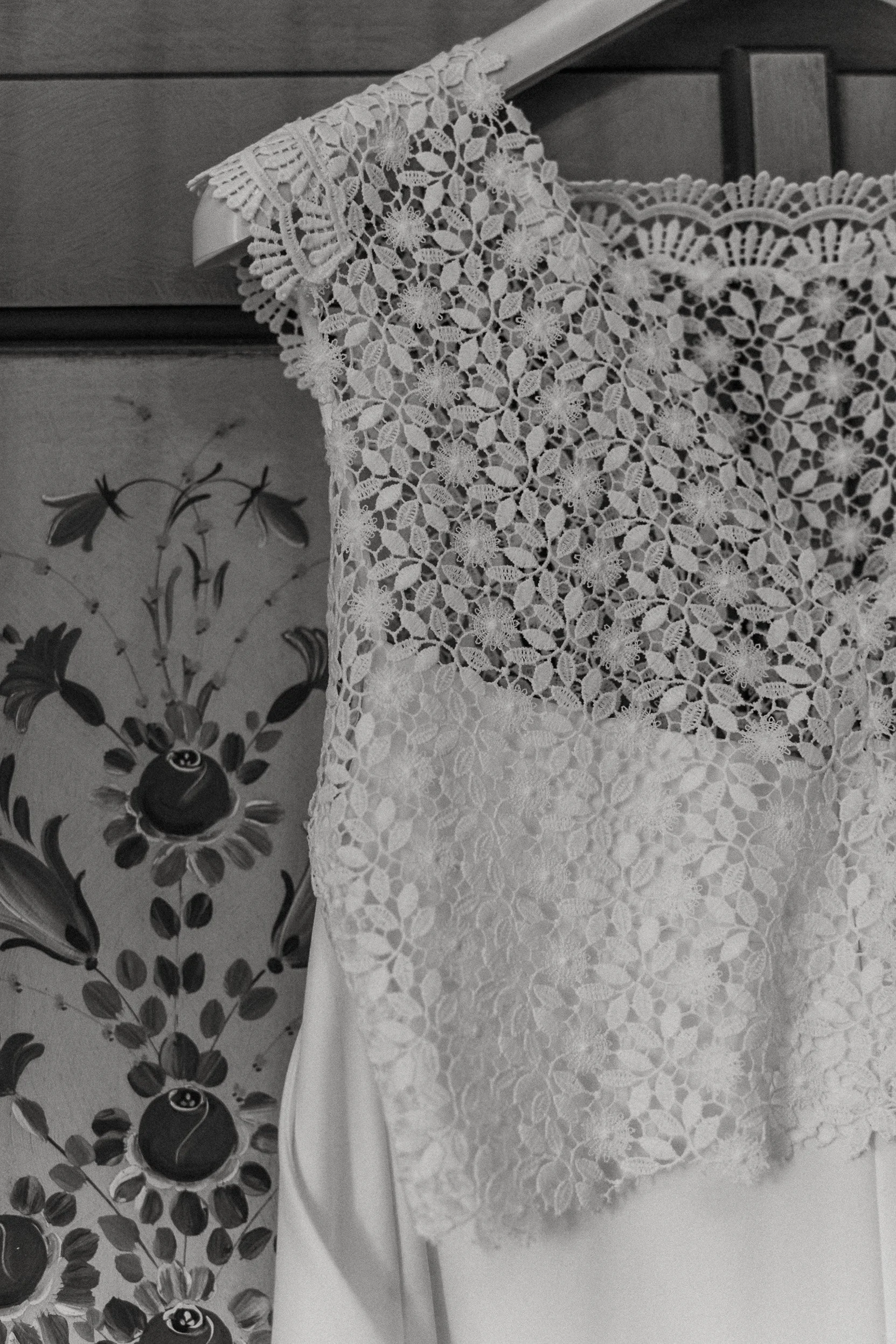 Vestito da sposa, tessuto di pizzo bianco con motivi floreali, appeso sopra uno sfondo con decorazioni di fiori stilizzati.
Brautkleid aus weißer Spitze mit floralen Mustern, aufgehängt vor einem Hintergrund mit stilisierten Blumendekorationen.