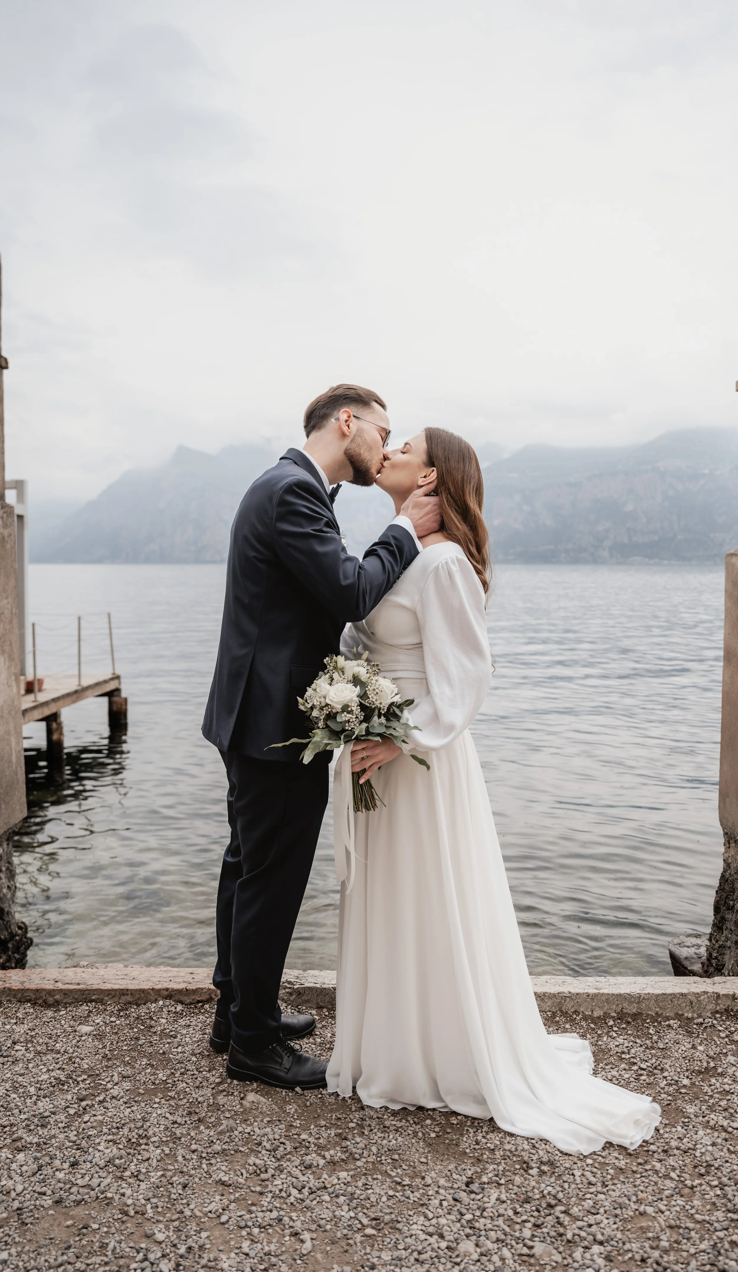 Ein Brautpaar, das sich vor einem See bei Nebel oder Wolken küsst, bei einer Hochzeit. Der Bräutigam trägt einen schwarzen Anzug und die Braut ein langes weißes Brautkleid, sie hält einen Blumenstrauß.
