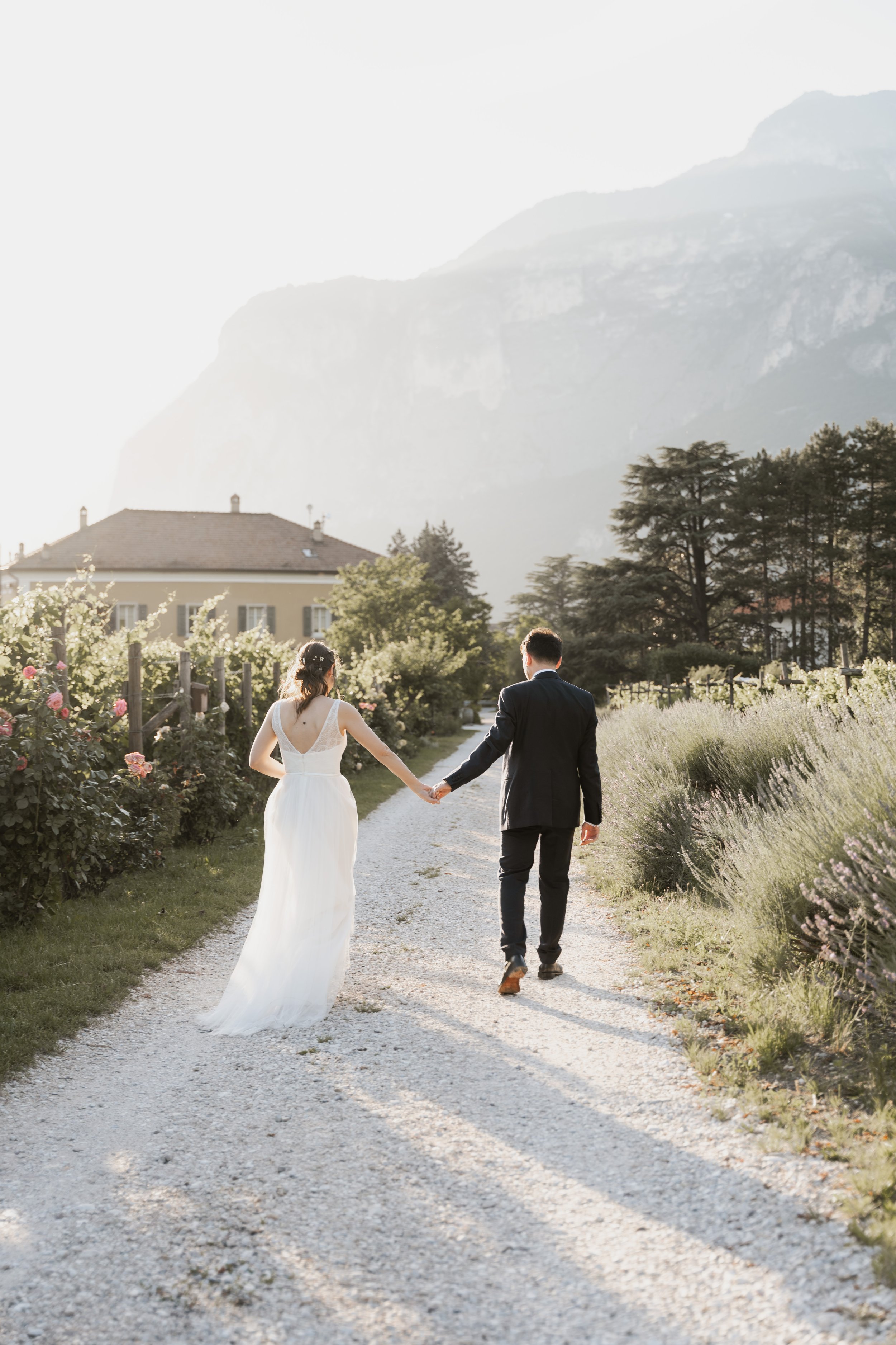 Ein junges Paar, ein Mann in einem schwarzen Anzug und eine Frau in einem weißen Hochzeitskleid, hält Händchen beim Spazierengehen auf einem Kiesweg in einem Garten mit Blumen und Bäumen, im Hintergrund Berge und ein Haus.
Hochzeitsfotografin Südtiro