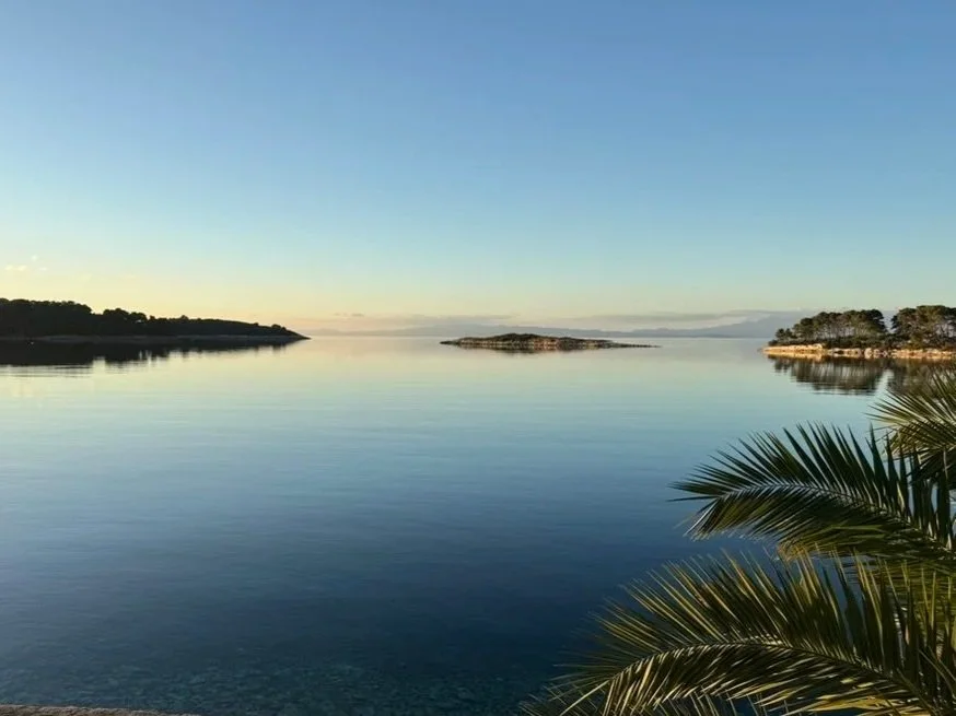 Calm body of water with small islands and tree-lined shores on either side, under a clear blue sky with distant mountains.