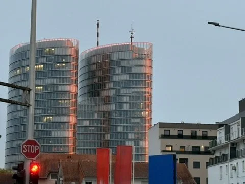 Two modern high-rise buildings with curved glass facades reflecting sunset colors, seen from the street with traffic lights and other buildings in the foreground.