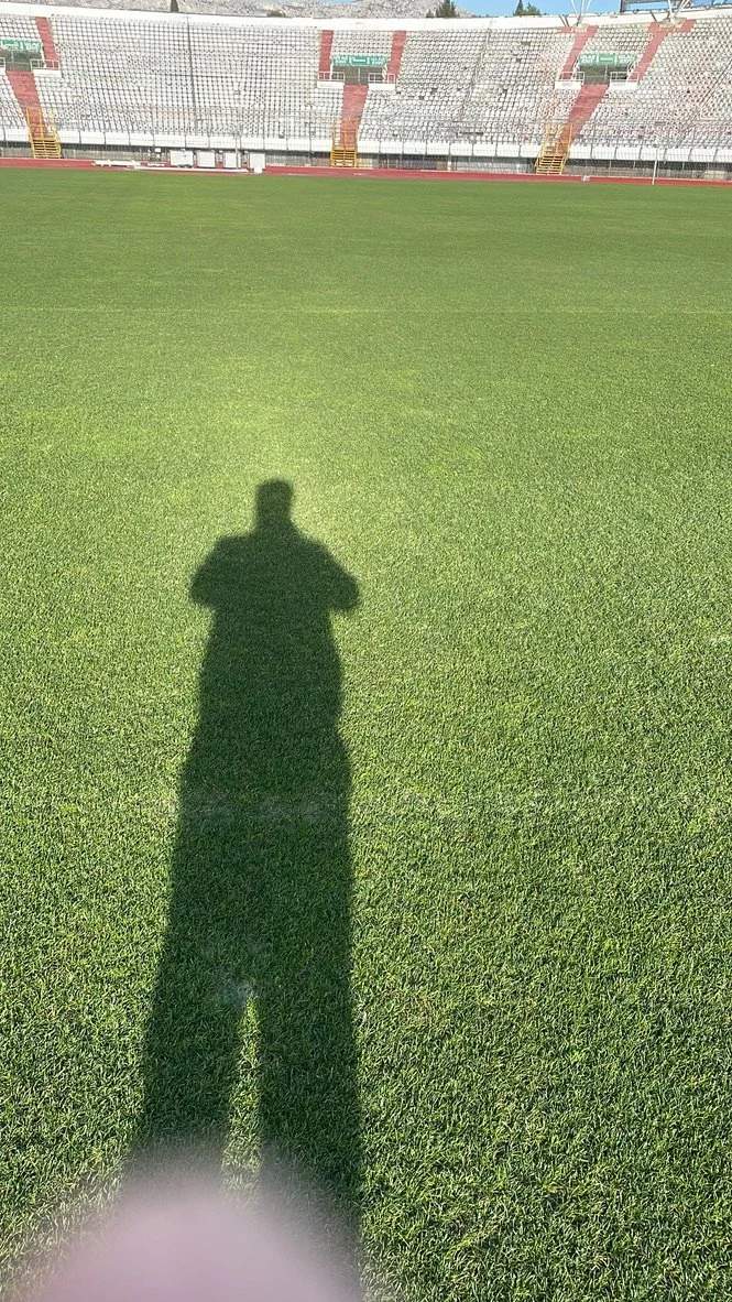 Empty sports stadium with a green field, red and gray seating, and the shadow of a person taking a photo, visible on the grass.