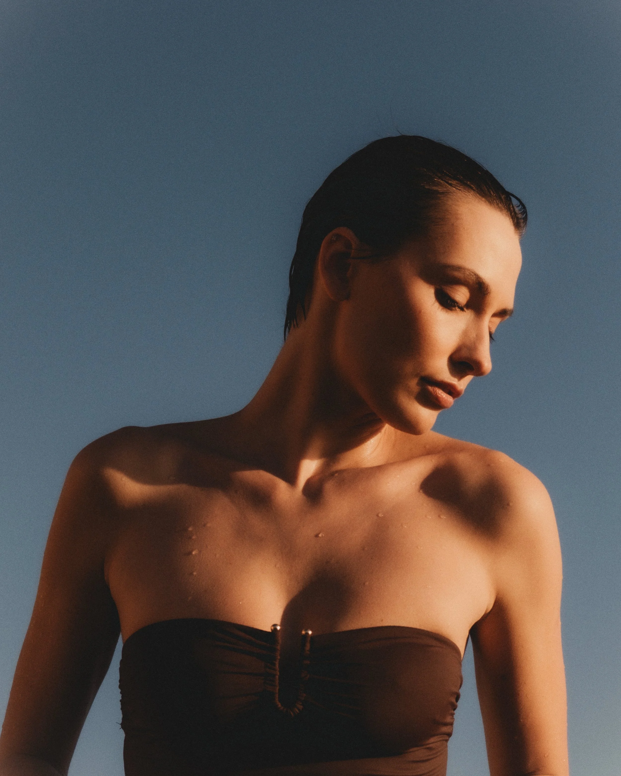 A woman with short wet hair and closed eyes, wearing a strapless top, set against a blue sky at sunset, with droplets of water on her shoulders.