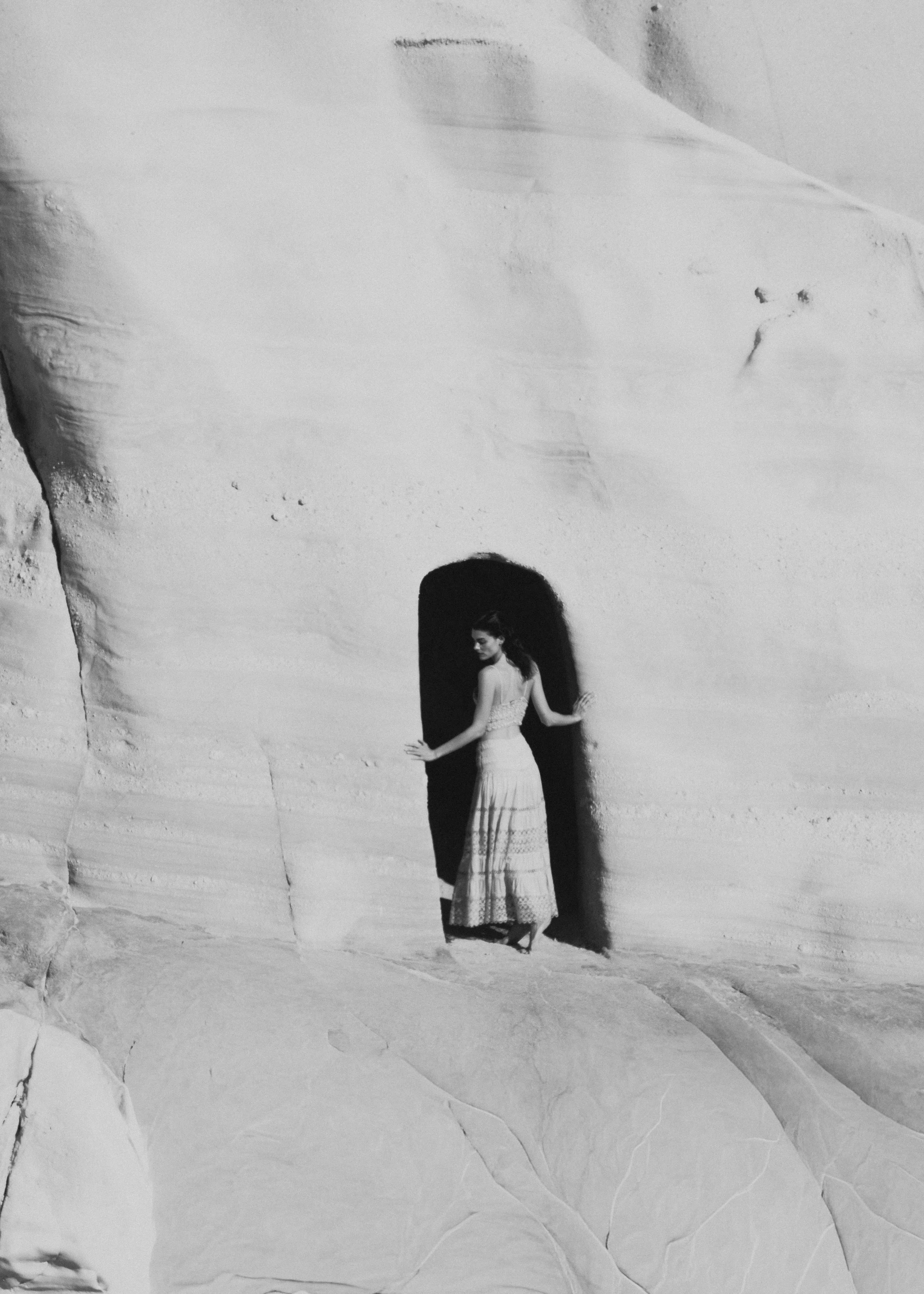 A woman in a long dress standing in front of a large shadow cast on a rocky wall, with her hand touching the wall.