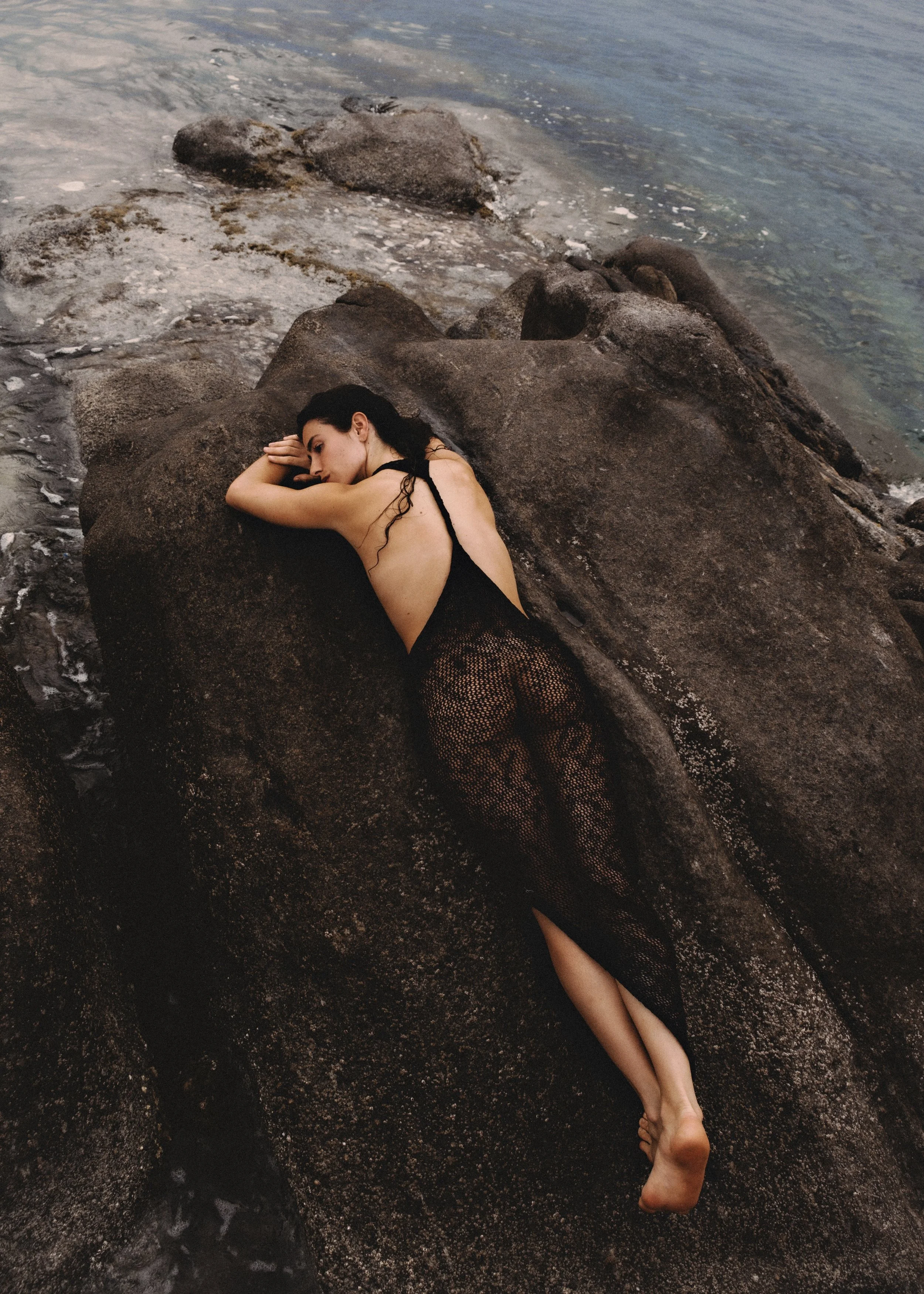 A woman with dark hair lying on her side on a large rock by the water, wearing a black dress with her eyes closed and arms resting on the rock.