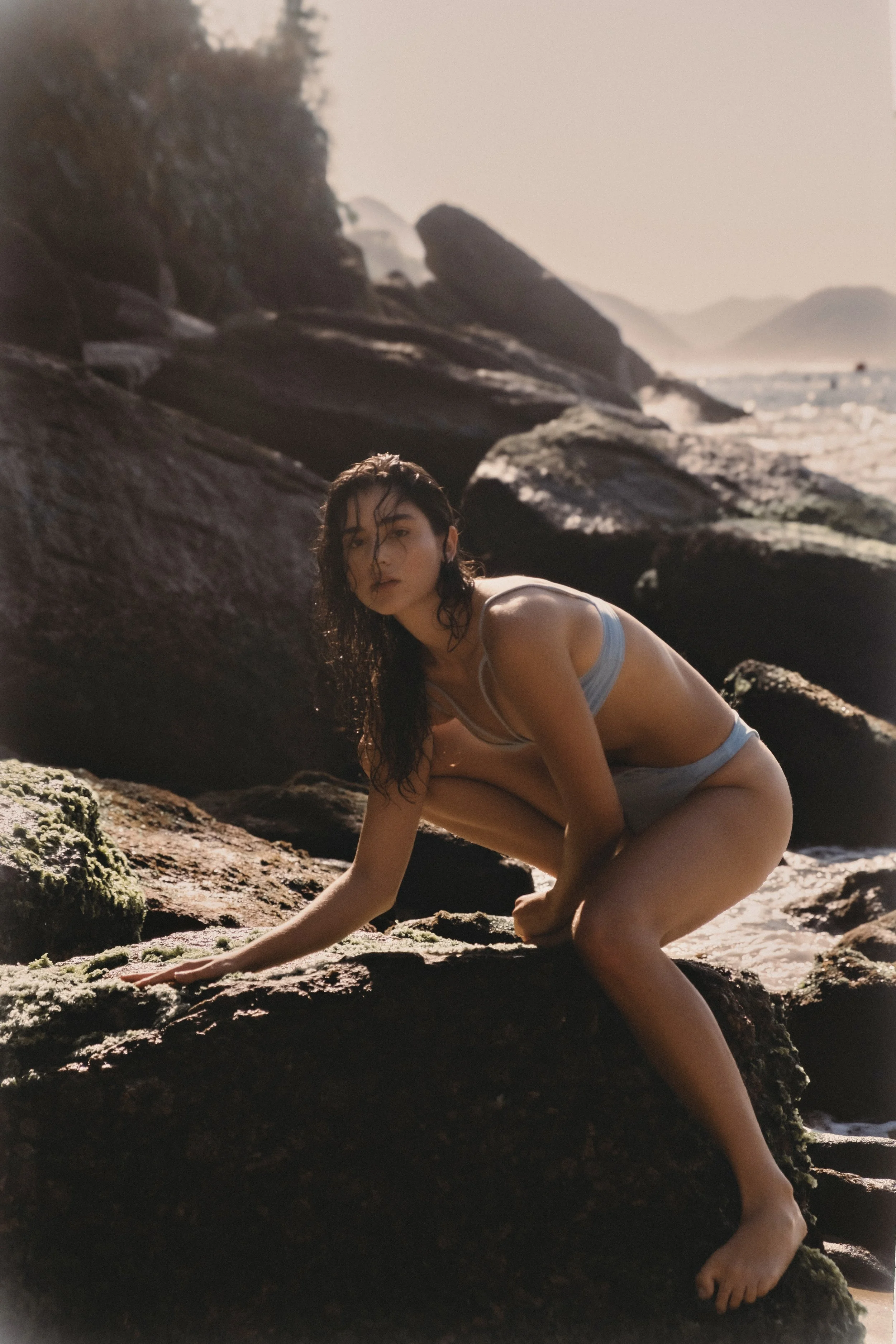 A woman with wet hair crouches on rocks at the beach, looking towards the camera, with the ocean and distant cliffs in the background.