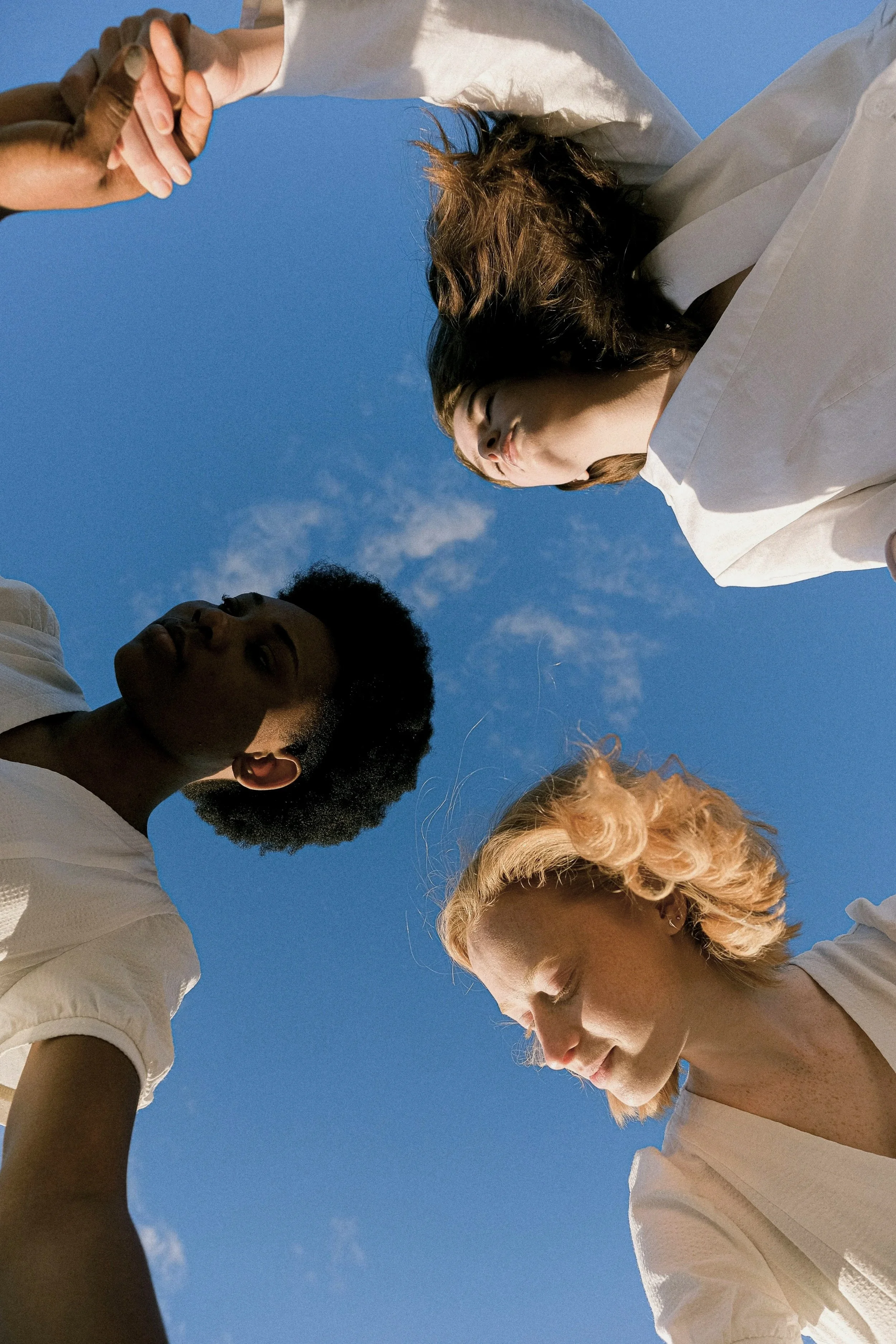 Group of diverse women standing in a circle outdoors, holding hands and looking inward, symbolizing collective identity, cultural connection, inclusion, and shared humanity.