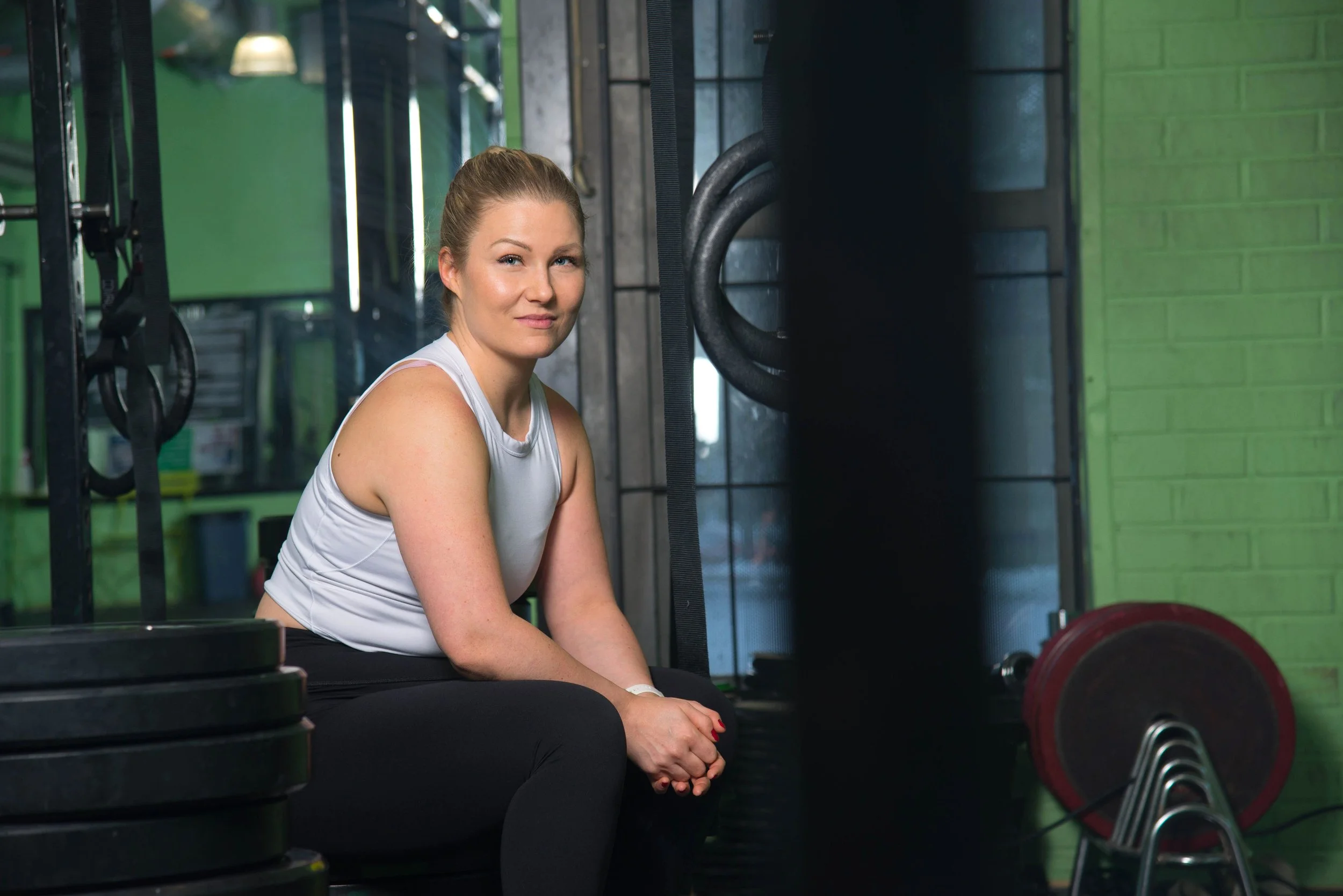 Nainen istuu kuntosalilla, ympärillä painonnostovälineitä ja kuntosalilaitteita