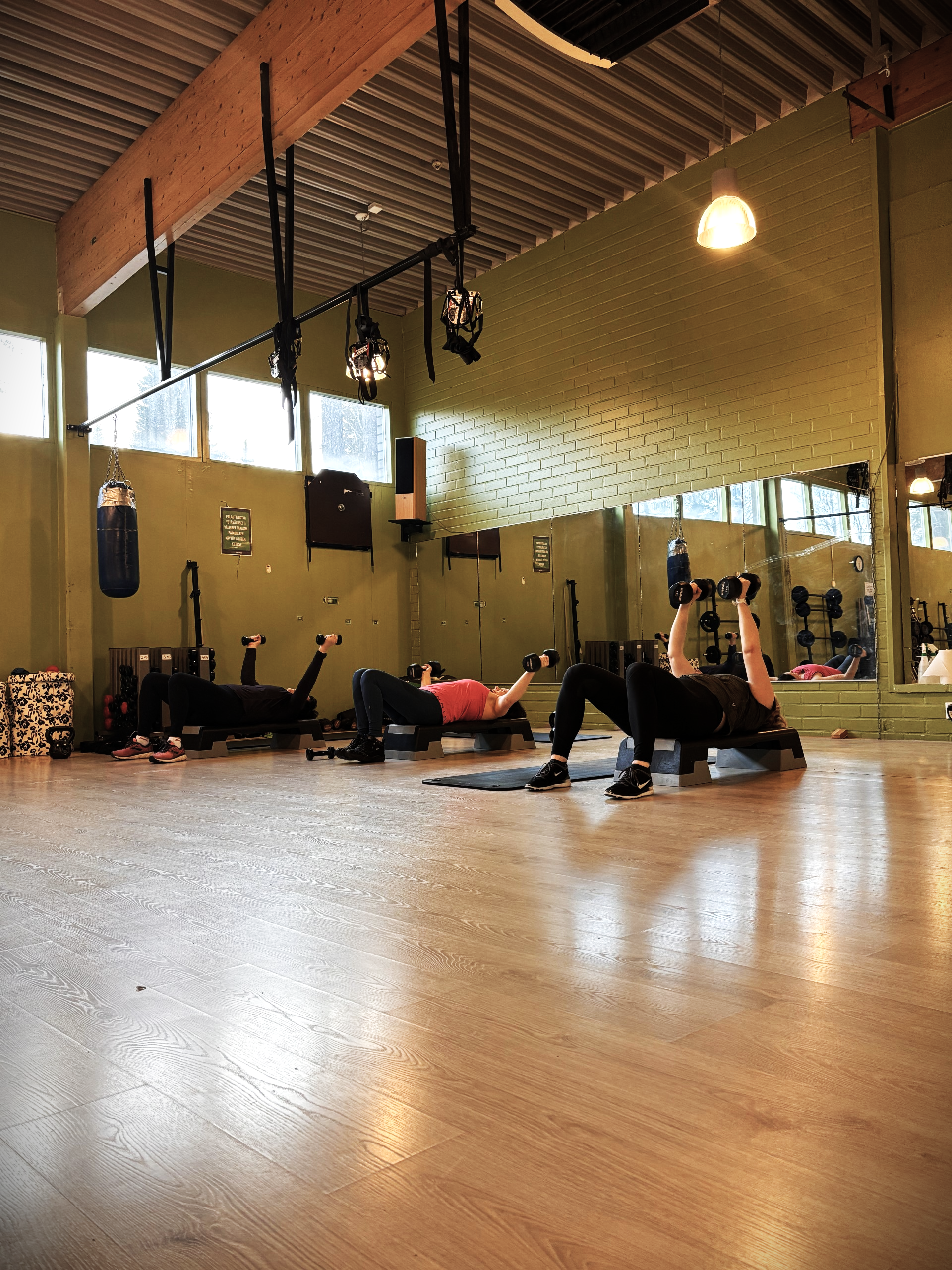 Naiskuva kuntosalilta, jossa kolme ihmistä harjoittelee treenimattojen päällä käsipainoilla.