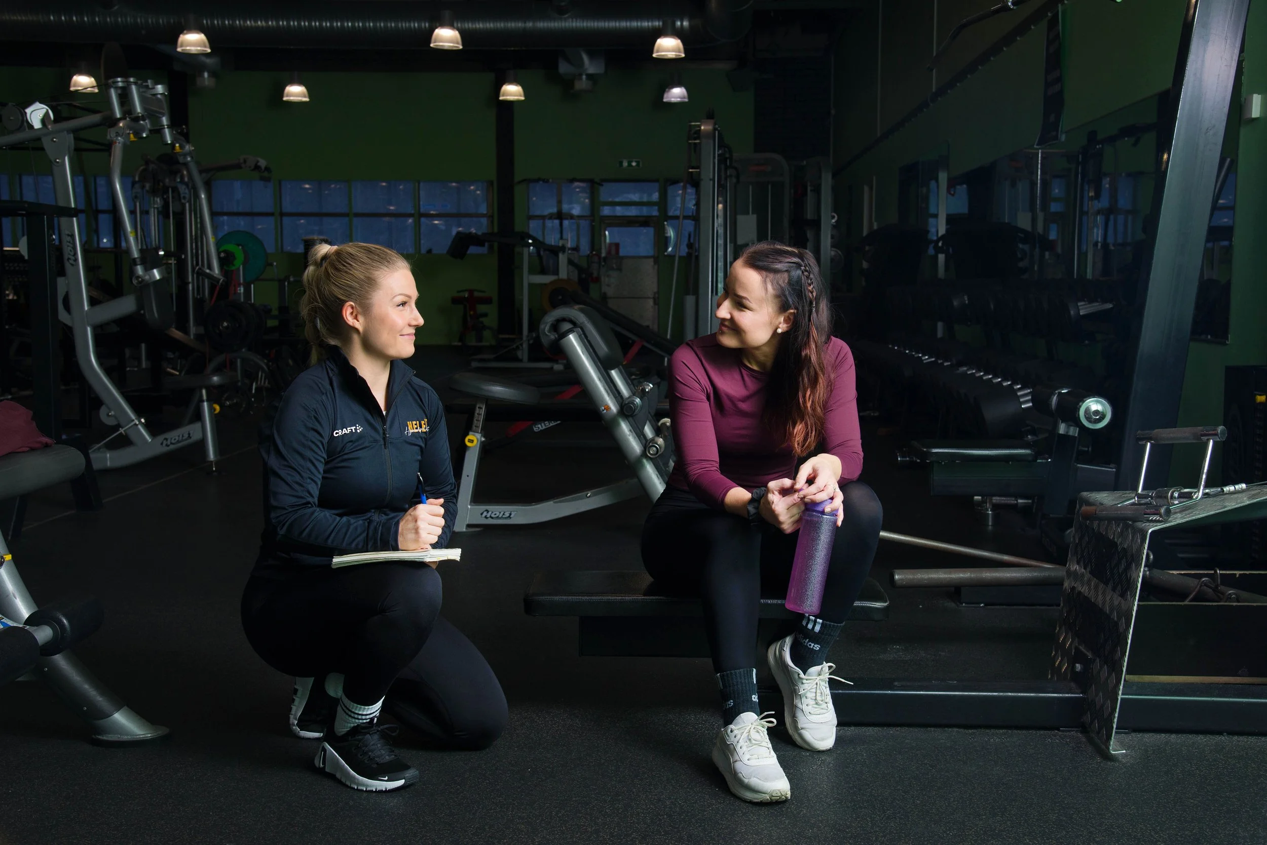Kaksi naista salitreenissä, toinen istuu penkillä ja toinen on polvillaan lattialla, keskustellessaan. Taustalla kuntosalilaitteita ja suuret ikkunat.