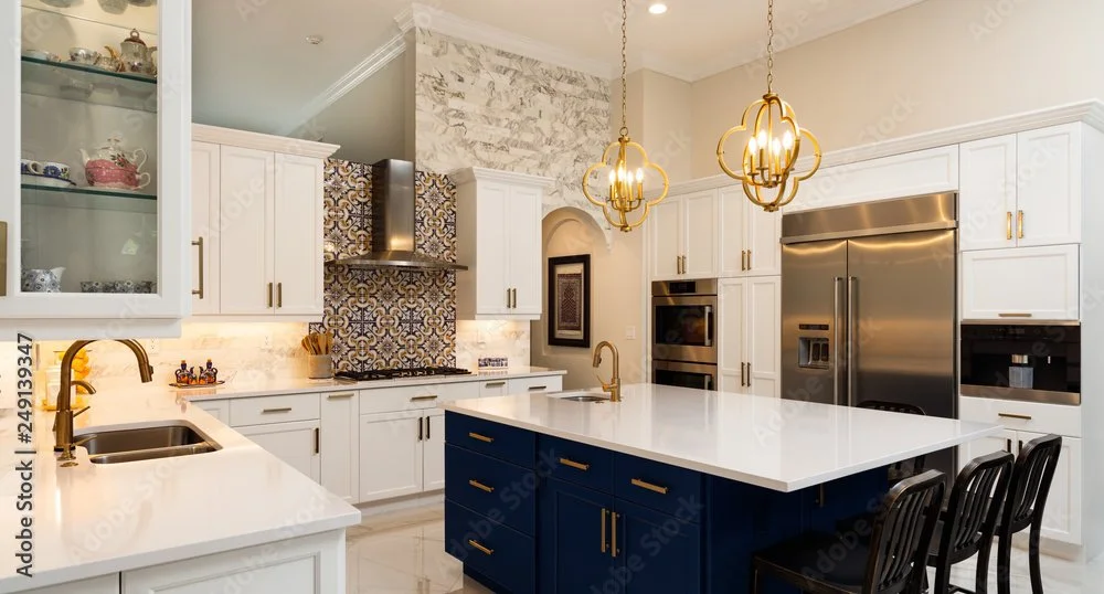 Modern kitchen with white cabinets, blue island with gold handles, marble countertop, stainless steel refrigerator, oven, and eating area with black chairs. Gold light fixtures hanging over the island and patterned backsplash behind the stove.