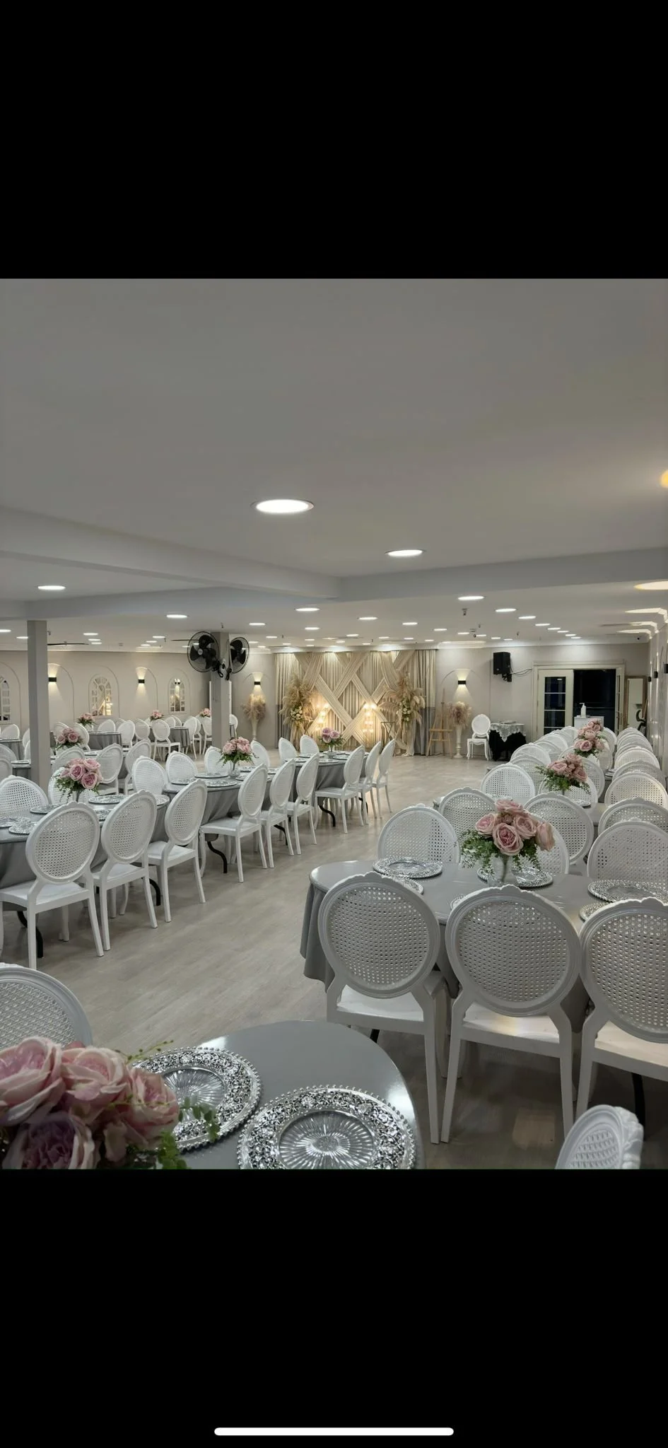 Düzenlenmiş nikah veya davet töreni için şık, beyaz ve gri masa ve sandalye dizilişi, ortasında çiçek aranjmanlarıyla süslenmiş. Arka planda dekoratif duvar ve aydınlatma bulunuyor.
