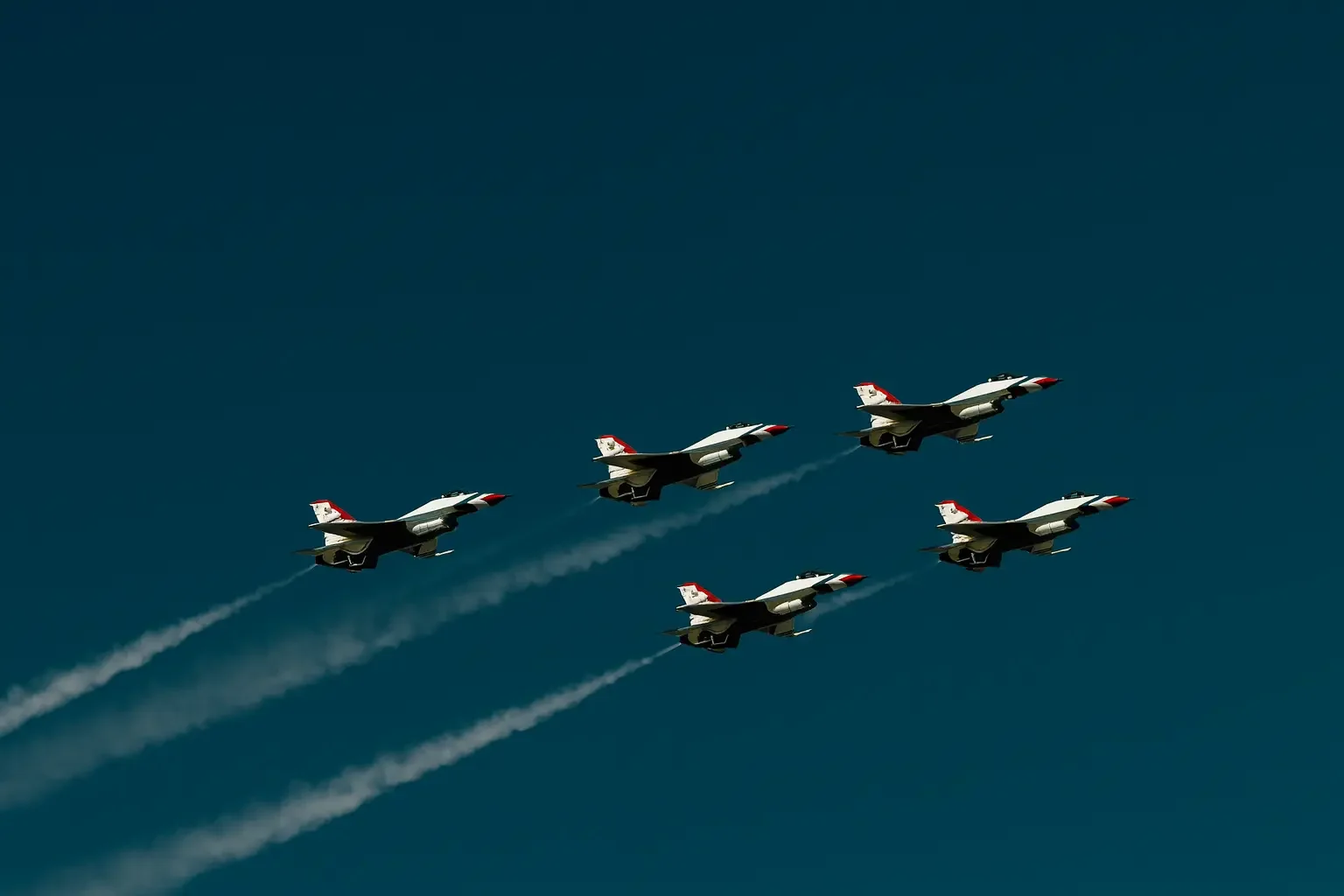 Fem jetfly som flyr i formasjon mot en klar blå himmel, og etterlater hvite kondensspor.