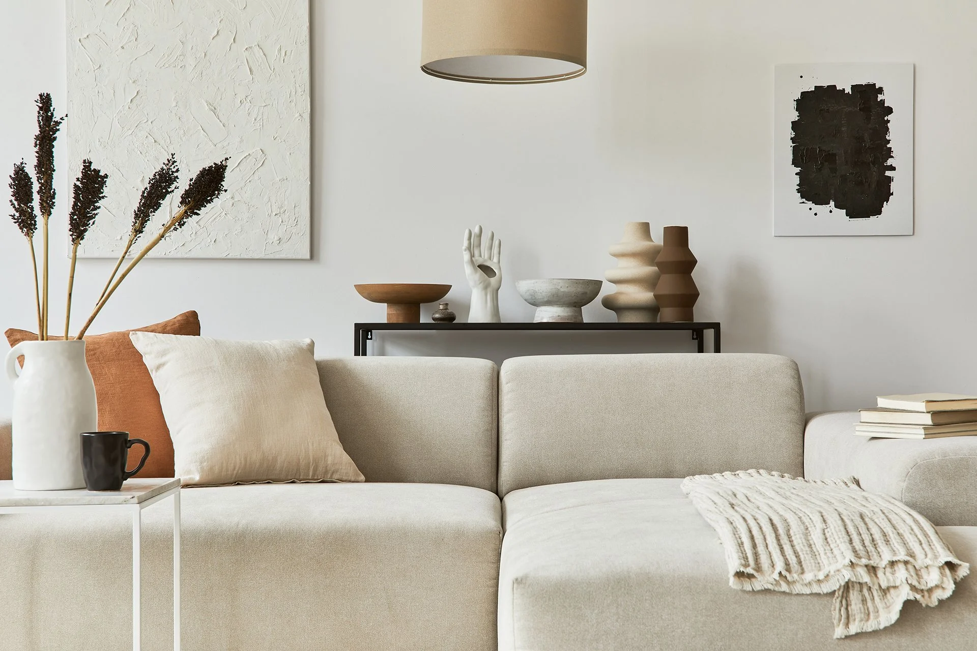 Kodikas olohuone, vaalea sohva, pöytä jossa kukkainen maljakko ja kahvikuppi, seinällä abstrakteja taideteoksia, koriste-esineitä hyllyllä, valkoinen ja beige sävymaailma.
