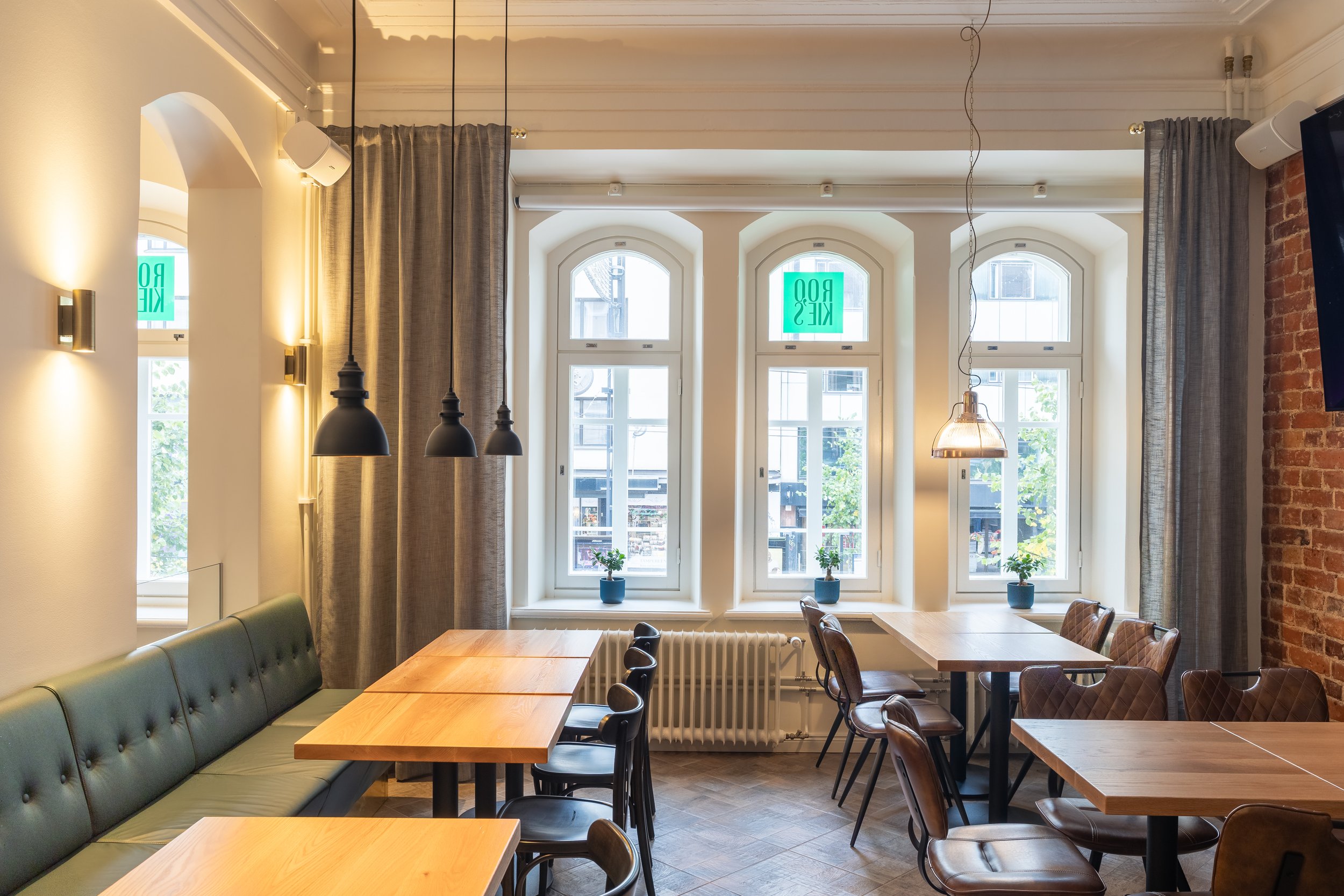 Sisätiloissa oleva ravintola tai kahvila, jossa on useita ruokapöytiä, ikkunat ja valoisaa luonnonvaloa, koriste-elementteinä kasvi ja laitteistoja, kuten valaisimia.