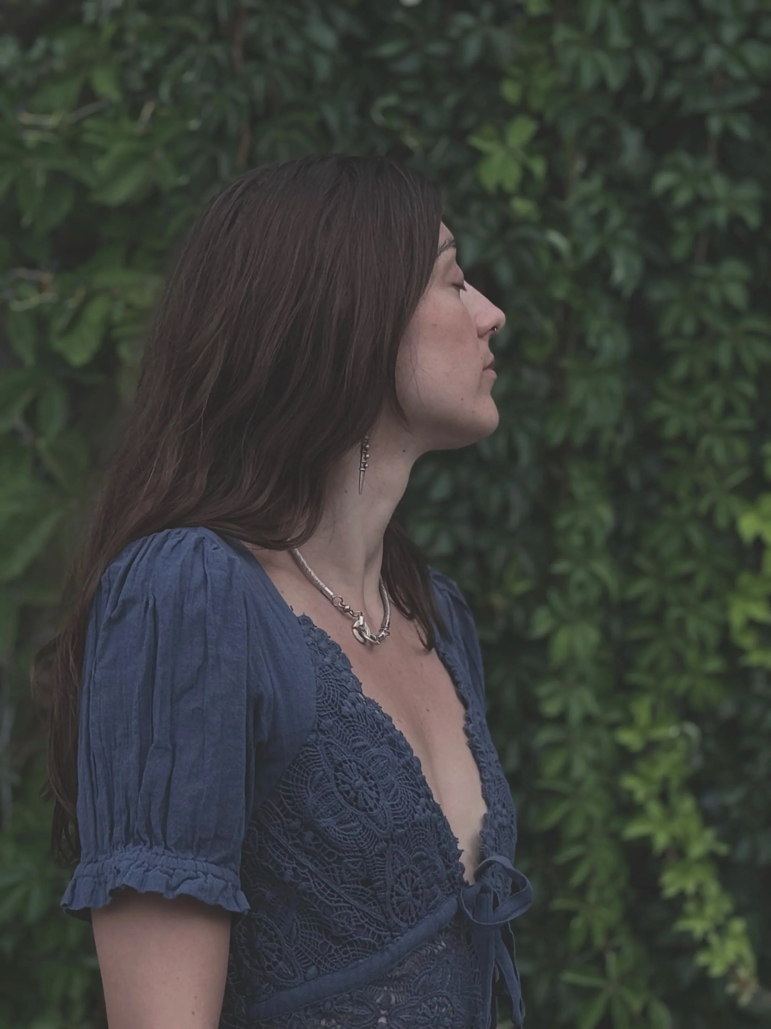 A woman with long brown hair standing outdoors in front of green foliage, wearing a blue lace dress, earrings, and a necklace, with her eyes closed and facing sideways.