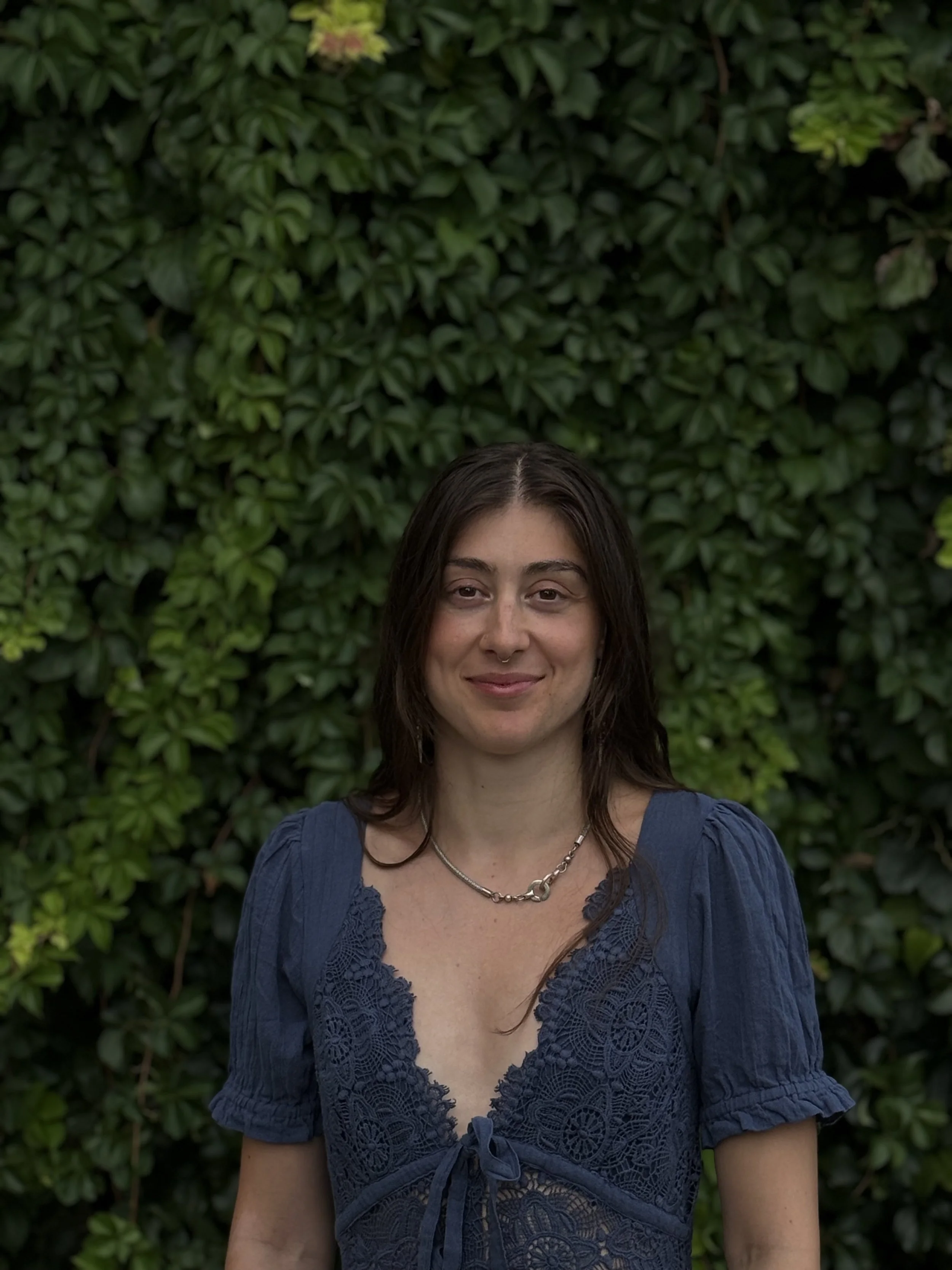 A woman with brown hair, wearing a blue lace dress and a silver necklace, standing outdoors in front of green foliage.