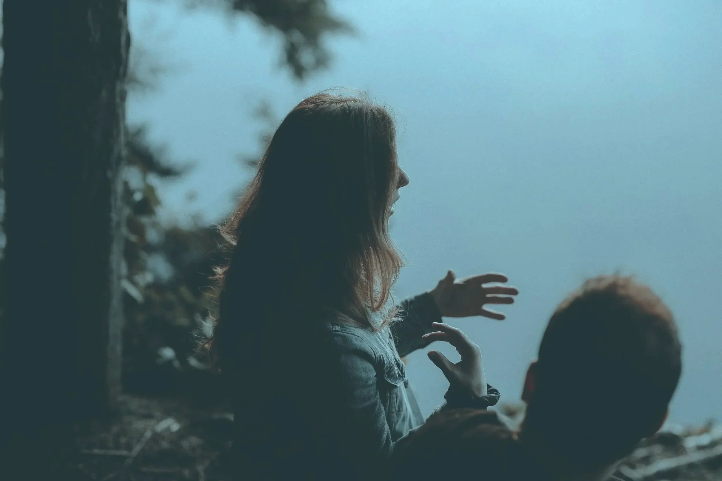 A woman with long hair talking & gesturing with her hands while standing outdoors near a tree, with a blurred person in the foreground and a misty or foggy background.