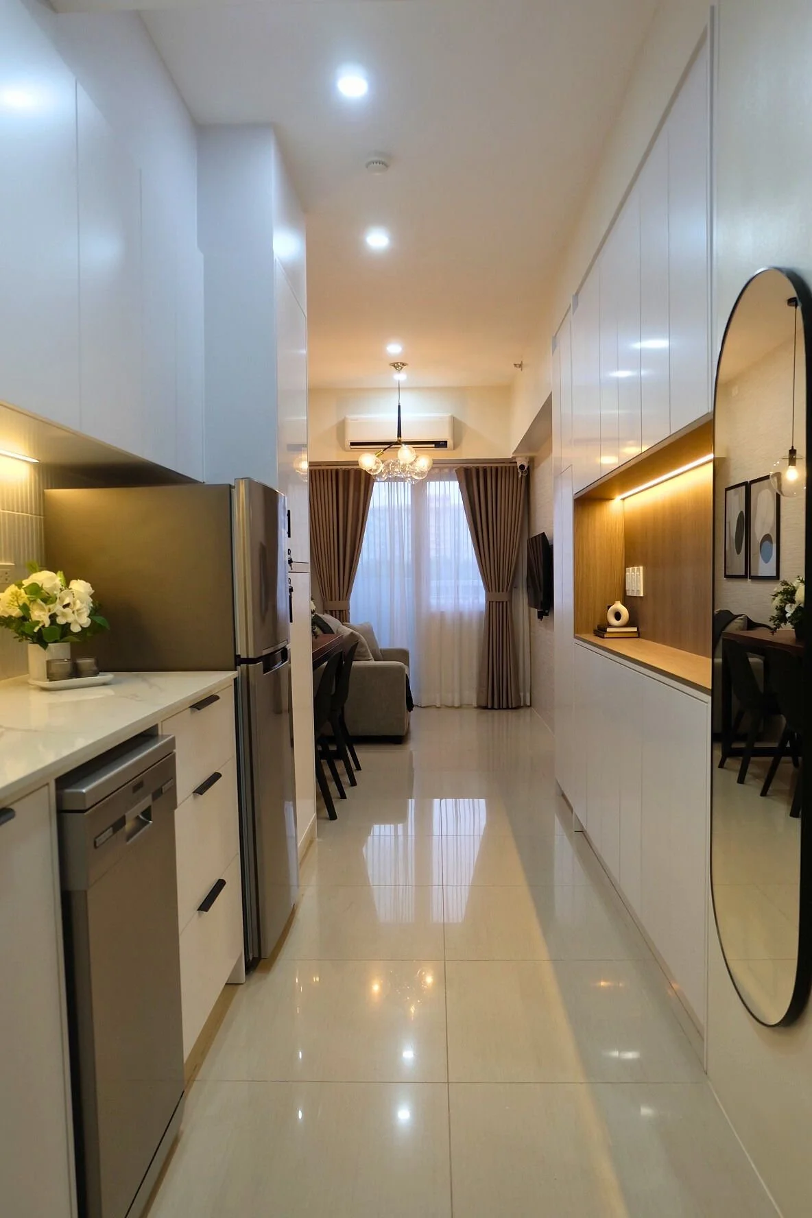 A modern, open-concept kitchen and living room area with white cabinetry, stainless steel appliances, a beige sofa, a dining table with chairs, and decorative artwork on the wall. Large windows with curtains allow natural light.