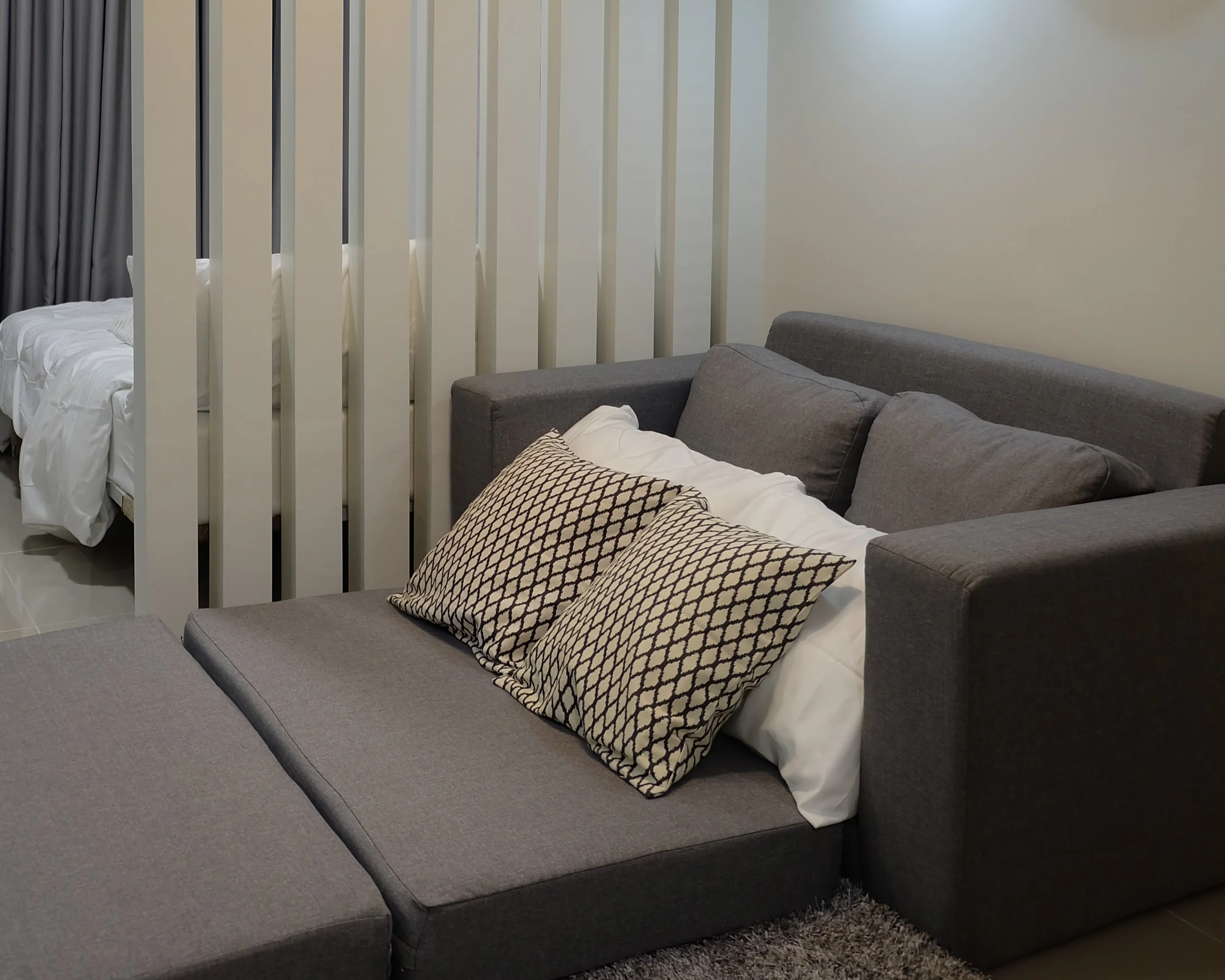 A gray sectional sofa with beige patterned pillows, a bed with white bedding, and a white room divider in a modern living space.