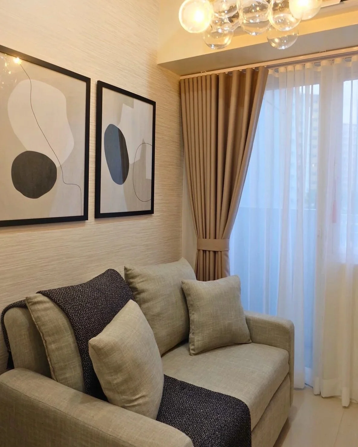 A living room corner with a beige sofa, decorative pillows, beige curtains, and abstract artwork on the wall.