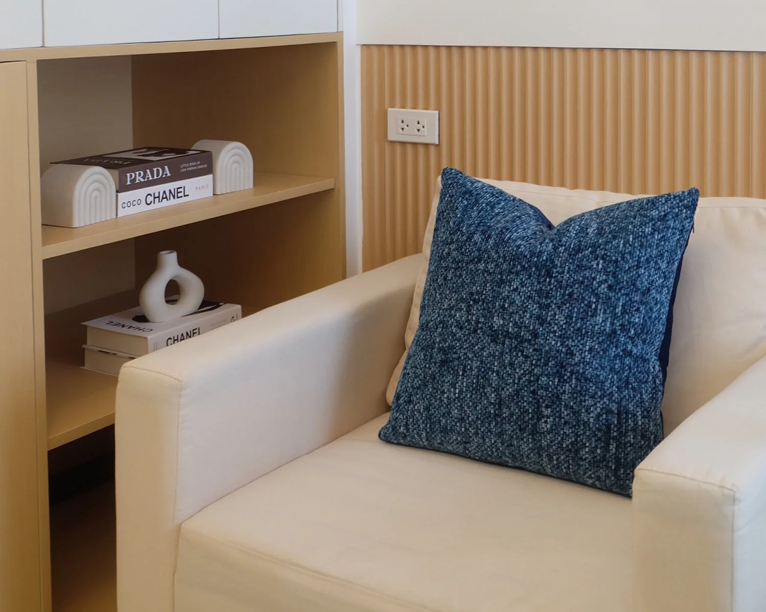A beige armchair with a large blue textured pillow placed on it. To the left, a wooden shelf with decorative books and a white ceramic vase, against a wall with built-in power outlets.