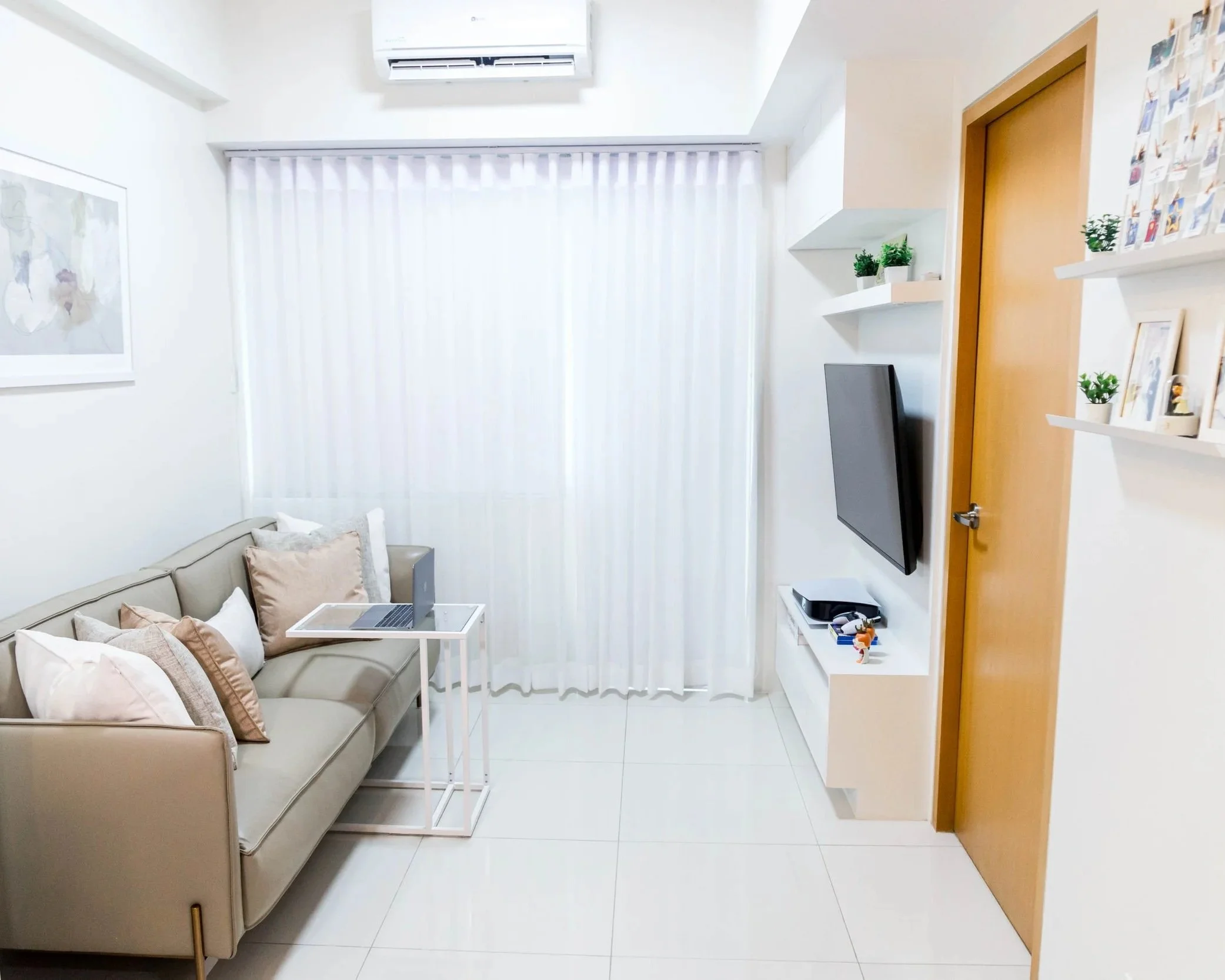 Bright modern living room with beige sofa, small white side table, wall-mounted TV, white shelving with decorative plants and photos, and curtains covering large window. Hardwood door is on the right.