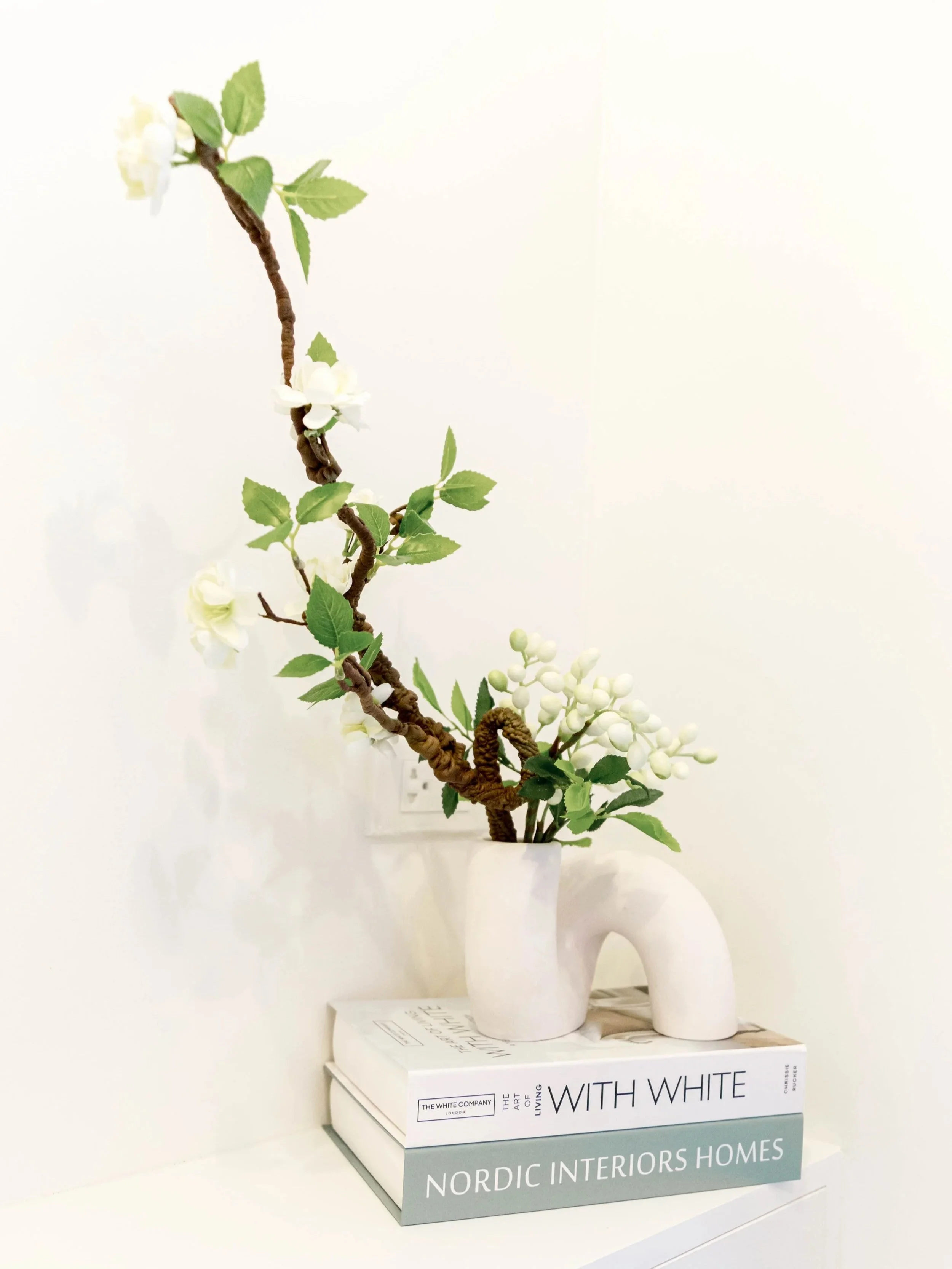 A white ceramic vase with an arch handle holding a flowering branch with green leaves and white blossoms. The vase is placed on top of two stacked books, one titled 'WITH WHITE' and the other 'NORDIC INTERIORS HOMES.' The background is plain white.