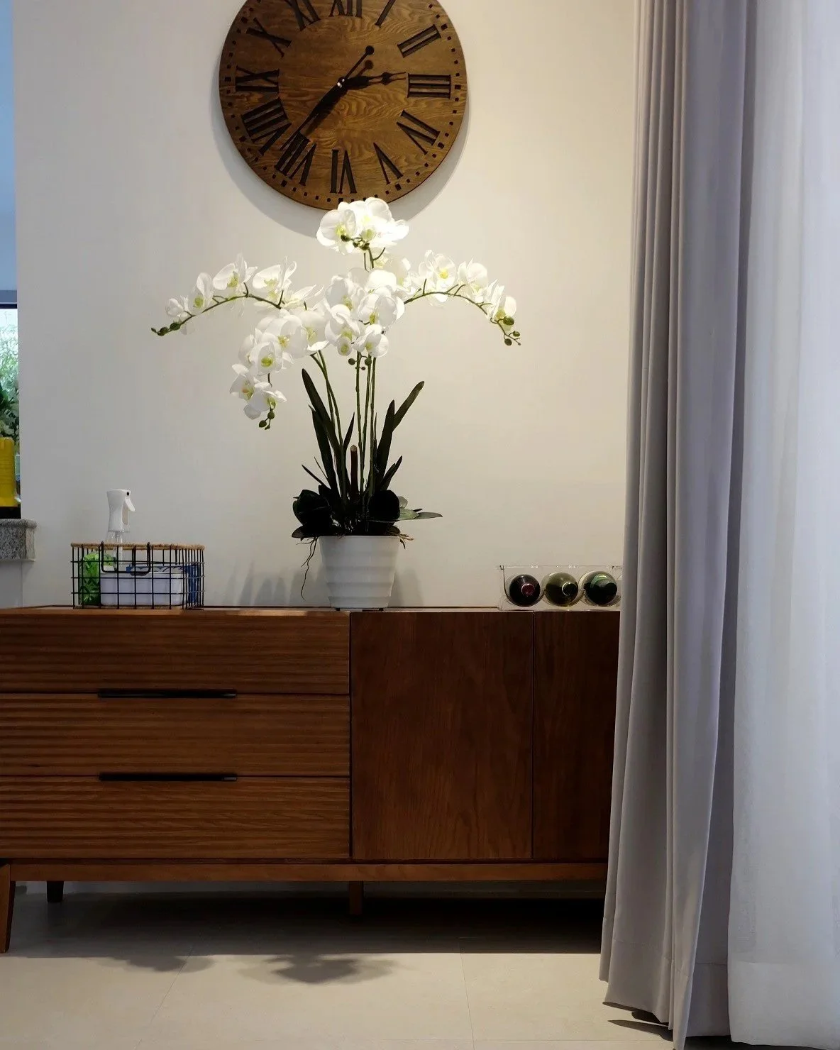 A wooden sideboard with a potted white orchid plant on top, a clock on the wall above, and decor items including spherical objects and a small basket, with curtains on the right side.