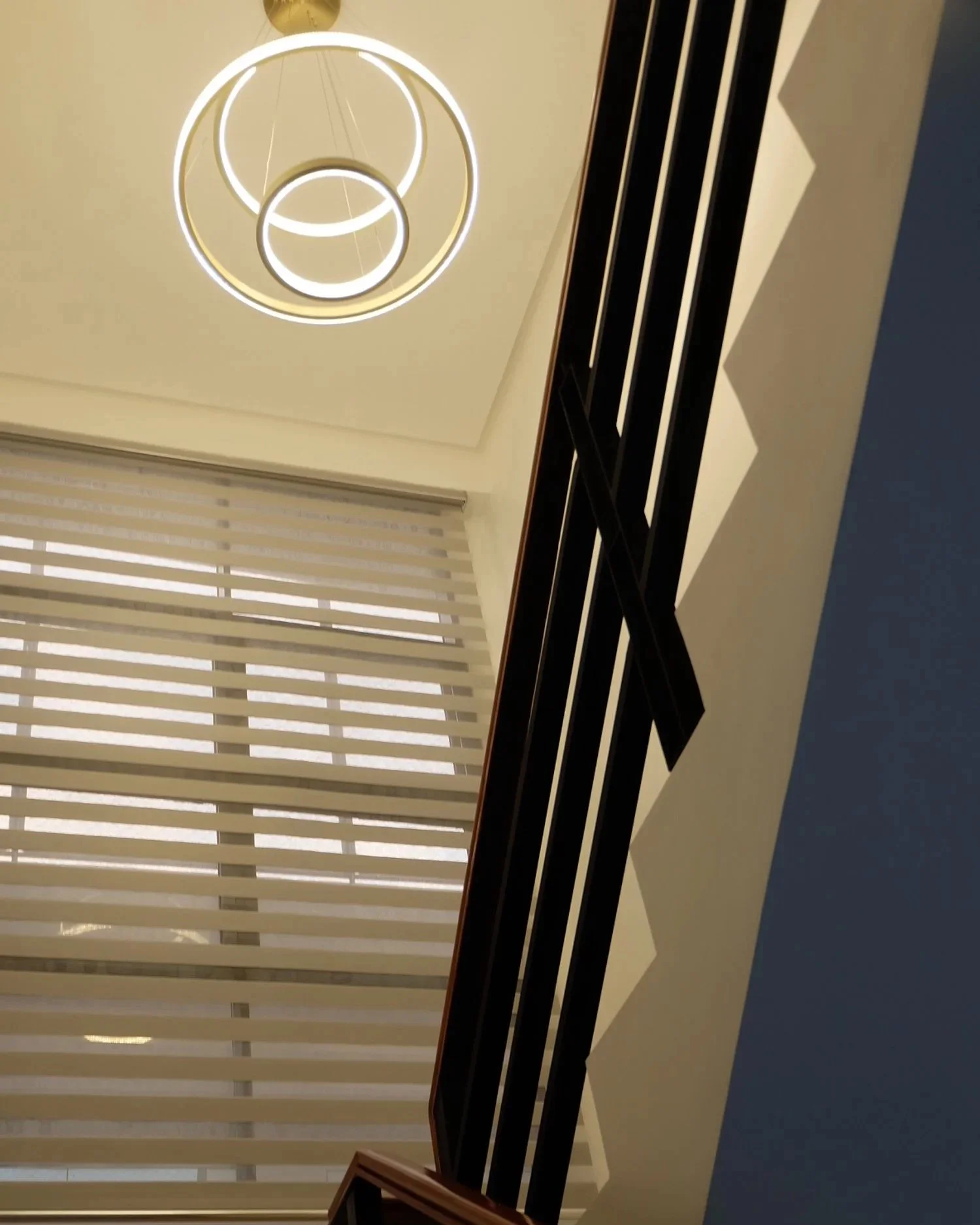 Modern interior view showing a staircase with black railing, a large window with beige blinds, and a ceiling light fixture with circular rings.