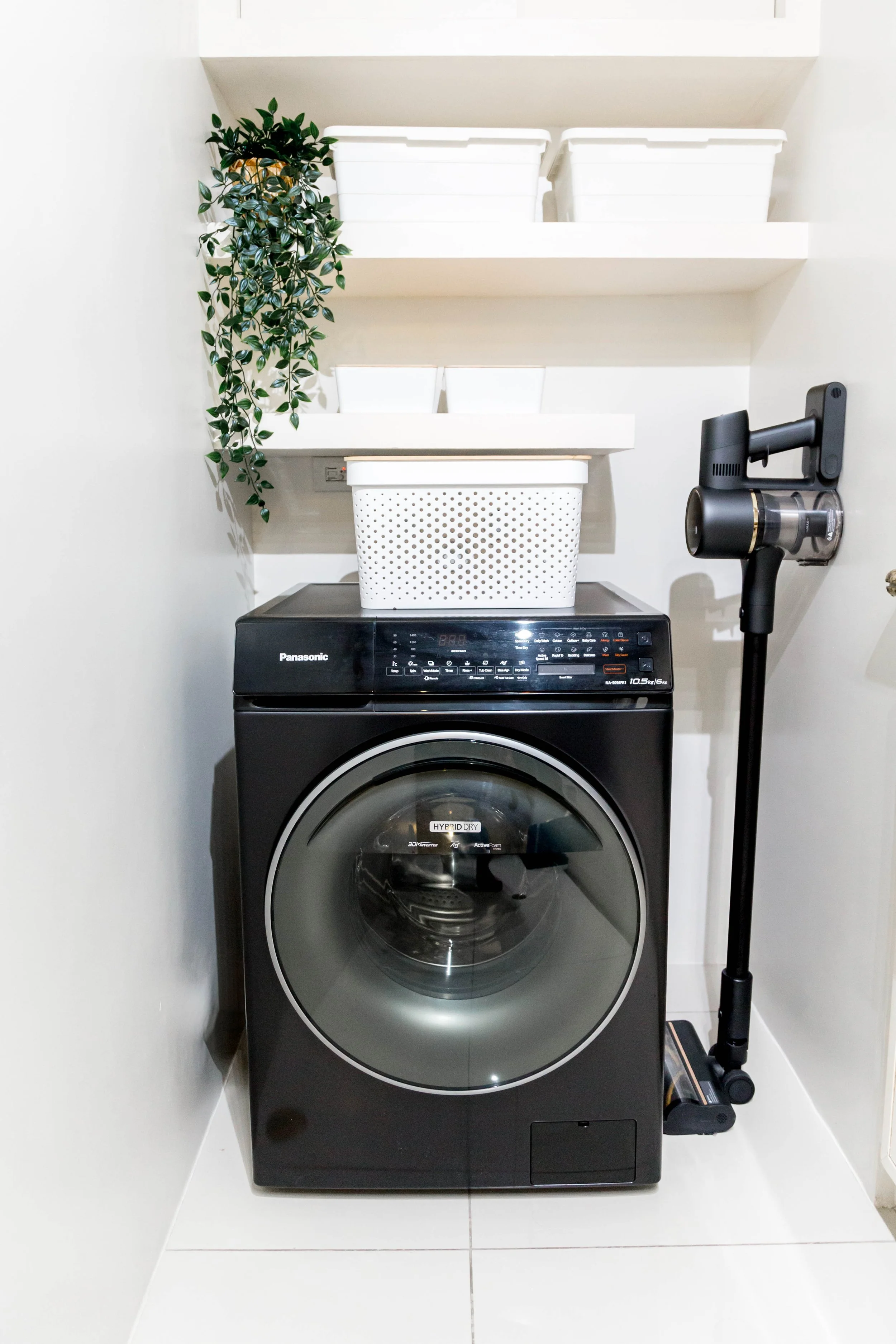 Front view of a black Panasonic front-loading washing machine with a digital display, positioned in a laundry area with white shelves holding white storage bins and a green leafy plant. A cordless vacuum cleaner is mounted on the wall to the right of